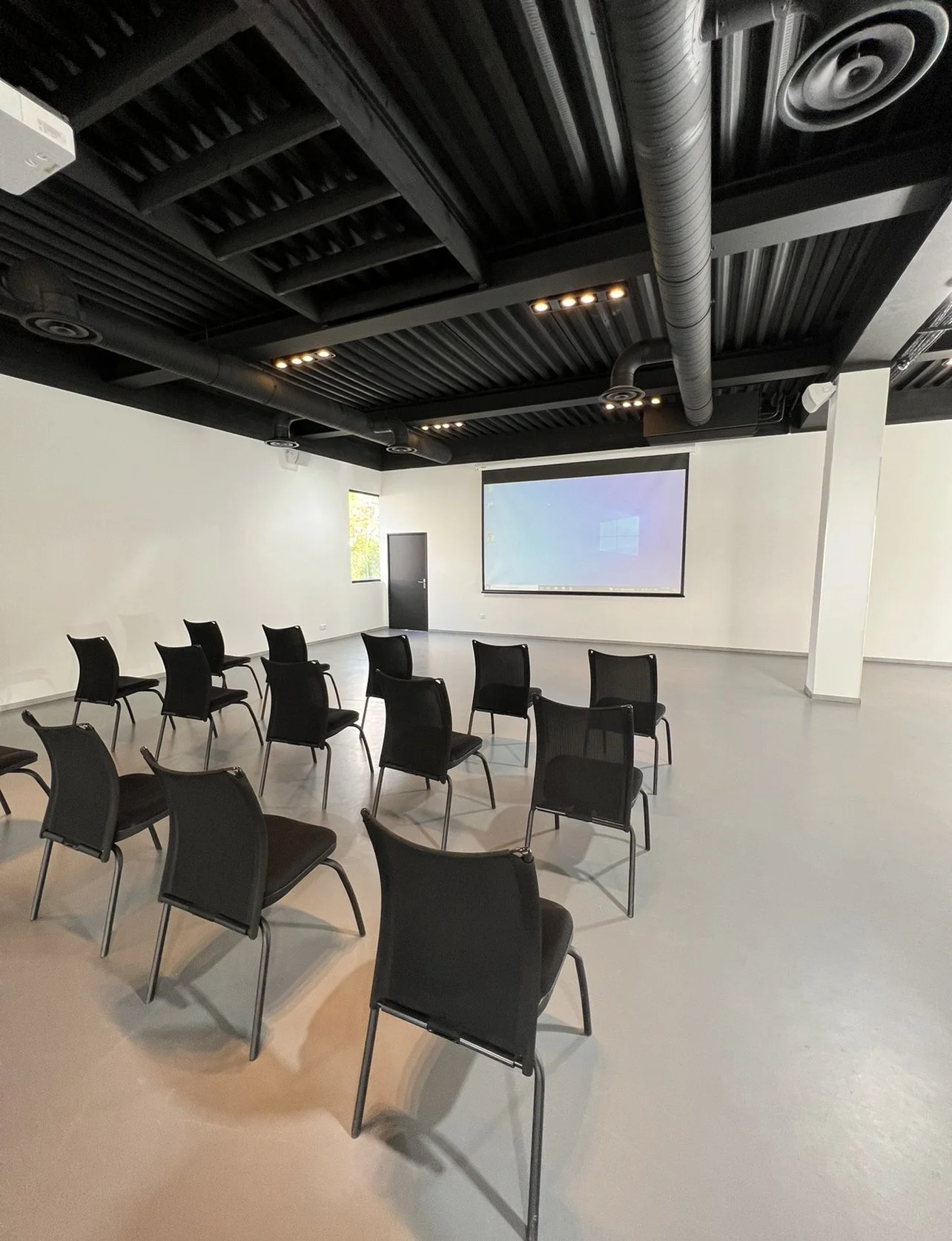 Une salle de conférence ou de projection avec plusieurs chaises noires disposées face à un grand écran de projection blanc. Le plafond est noir avec des conduits visibles et des lumières encastrées.