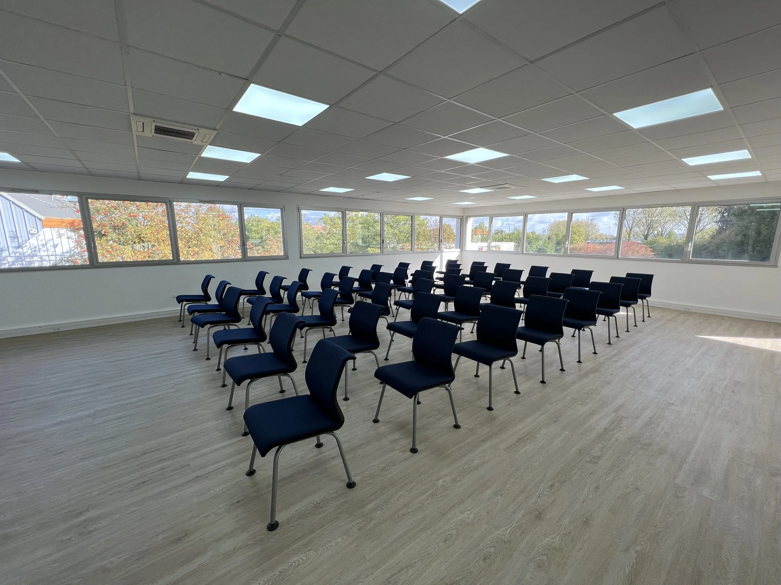 Salle de réunion ou conférence avec plusieurs rangées de chaises bleues disposées à l'intérieur, vue sur des fenêtres avec un paysage extérieur, plafond suspendu avec éclairages intégrés.