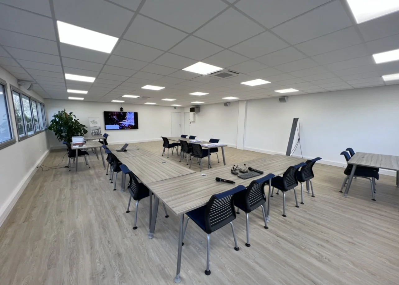 Salle de réunion équipée de tables en U avec des chaises, un grand écran mural, une plante verte à gauche, et des équipements électroniques au centre.