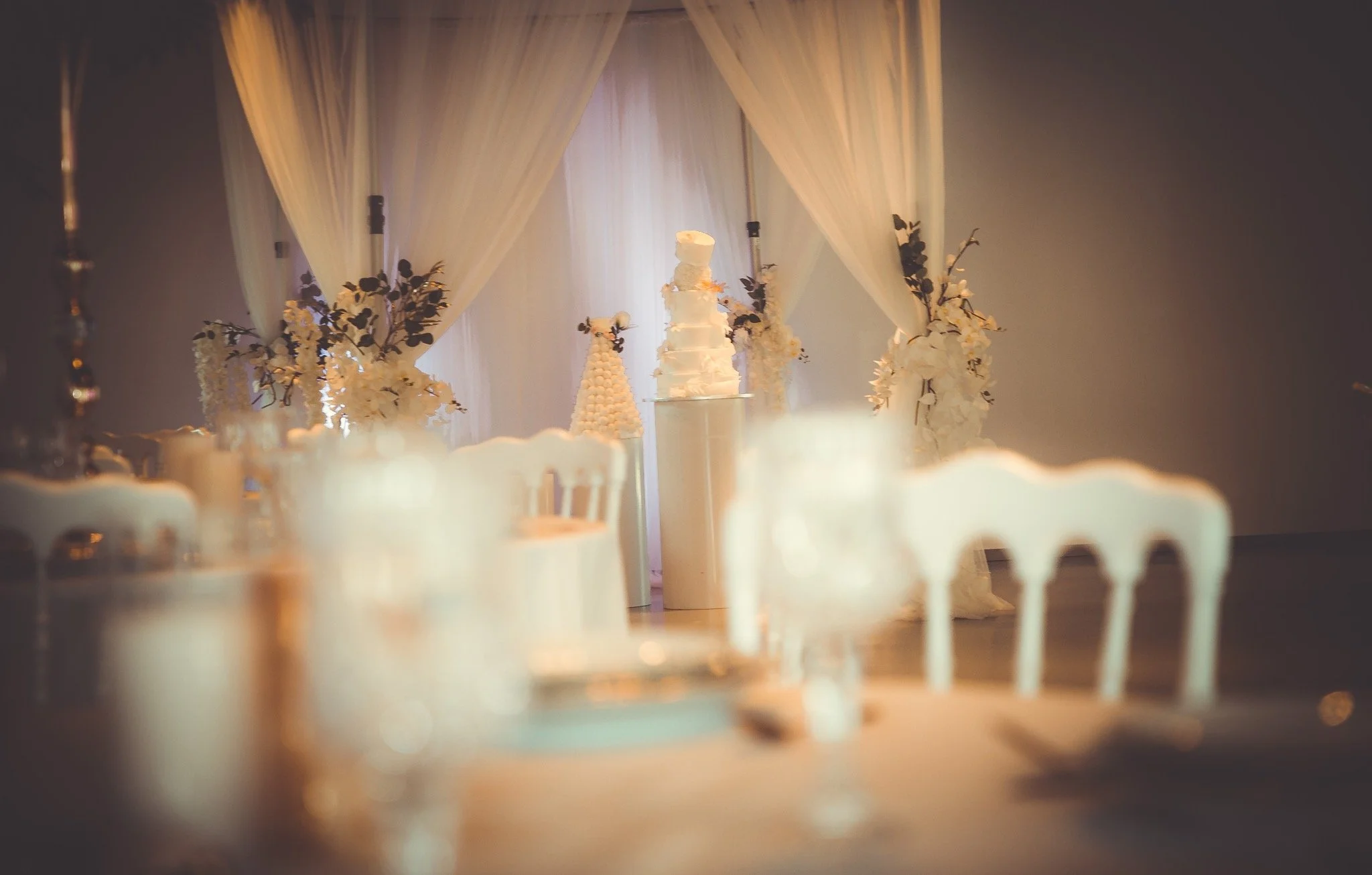 Une décoration de mariage élégante avec un gâteau blanc à plusieurs étages au centre, entourée de fleurs blanches et de bougies, avec un décor en tissu léger et des rideaux blancs à l'arrière.