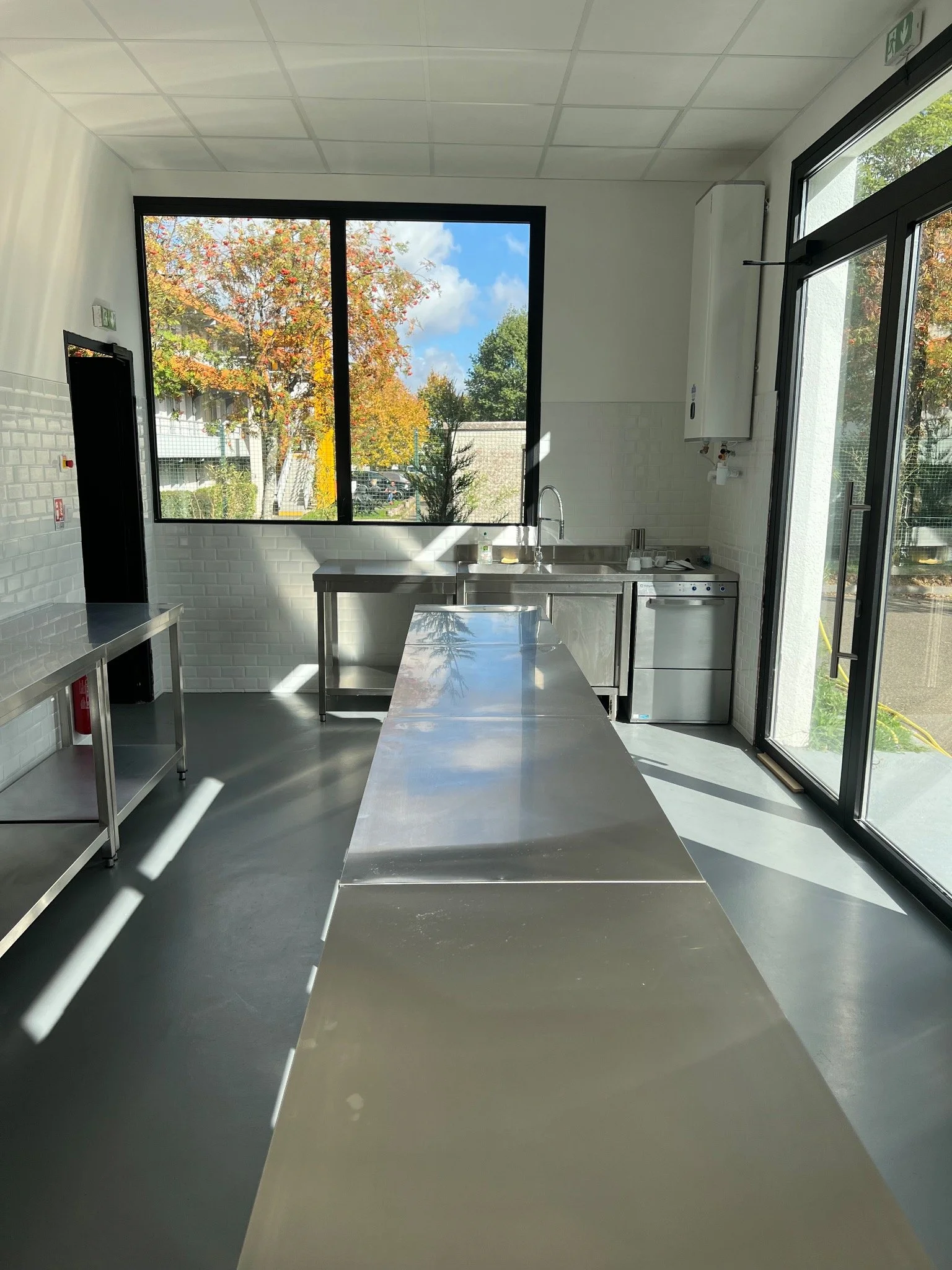 Cuisine lumineuse avec surfaces en acier inoxydable, grande fenêtre avec vue sur des arbres colorés d'automne, porte coulissante en verre.