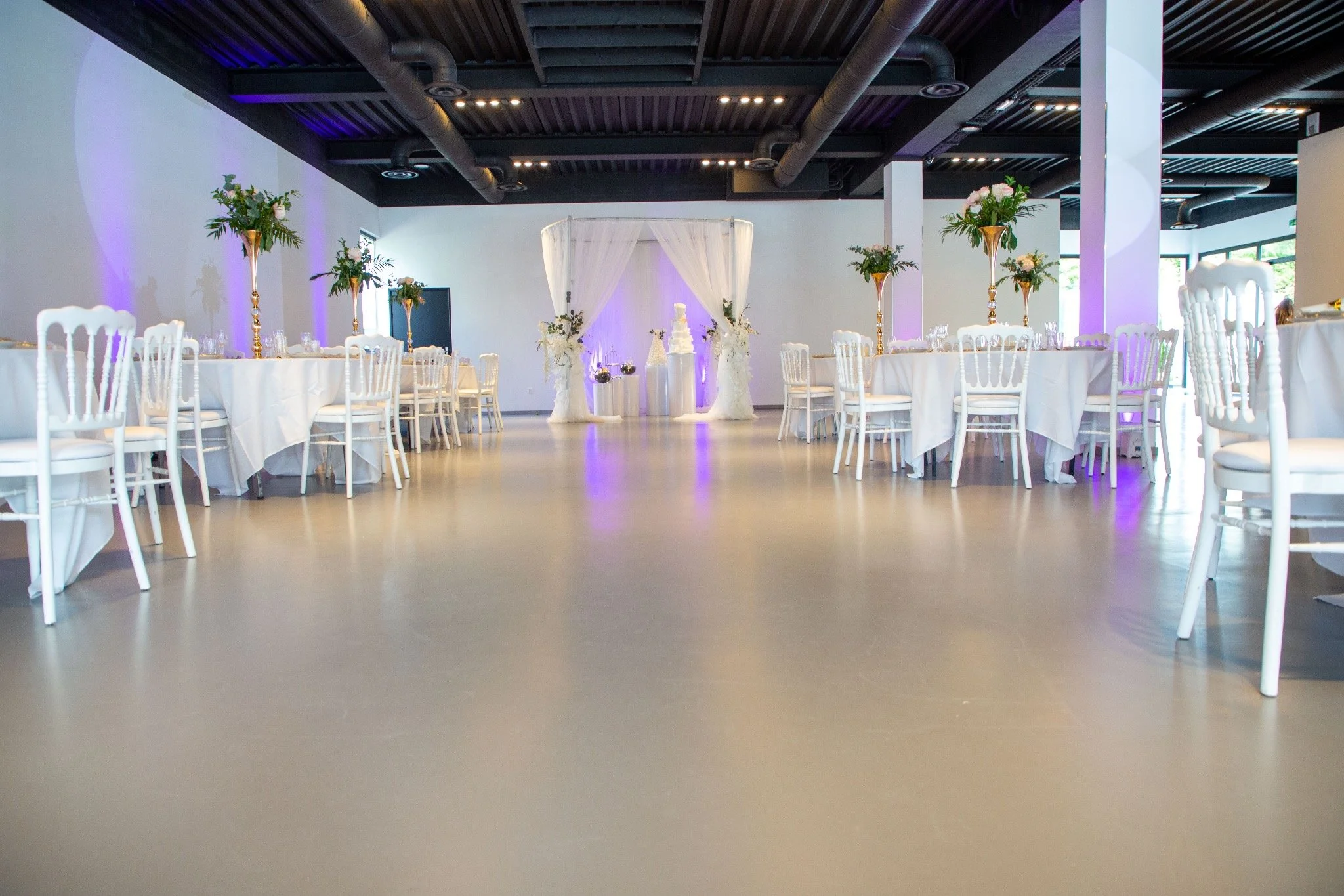 Salle de réception élégante avec tables rondes drapées de blanc, chaises blanches, décor floral en hauteur, et une zone centrale pour la cérémonie avec une toile blanche et des fleurs, éclairée de lumières douces