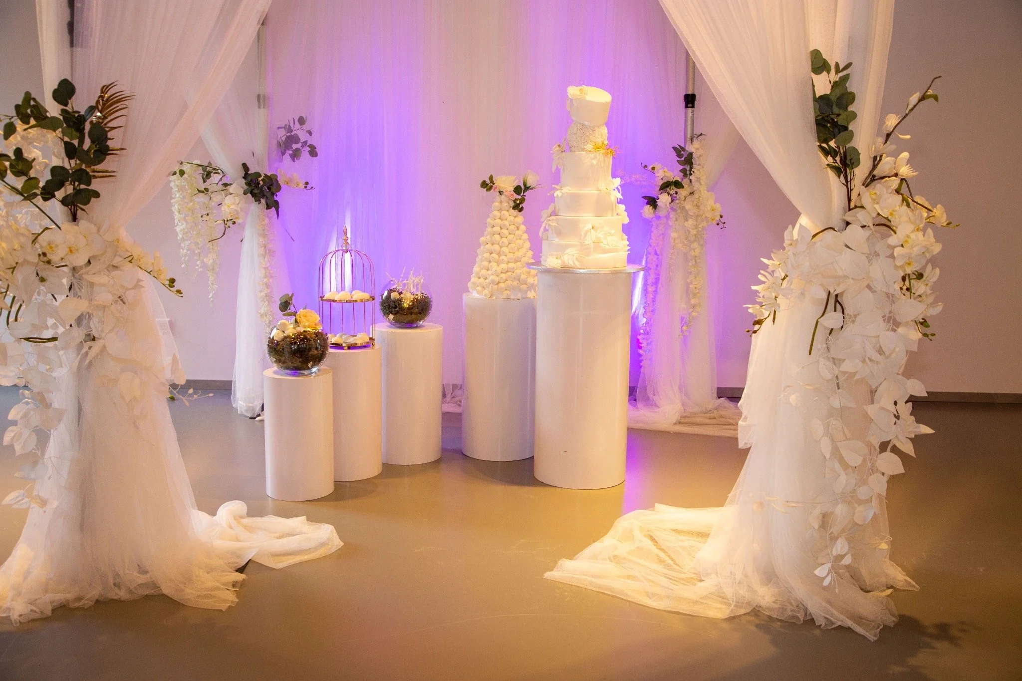 Décor de mariage avec un gâteau de plusieurs étages, entouré de fleurs blanches et de lanternes blanches sur des piédestaux, avec un fond en tissu blanc et un éclairage violet.