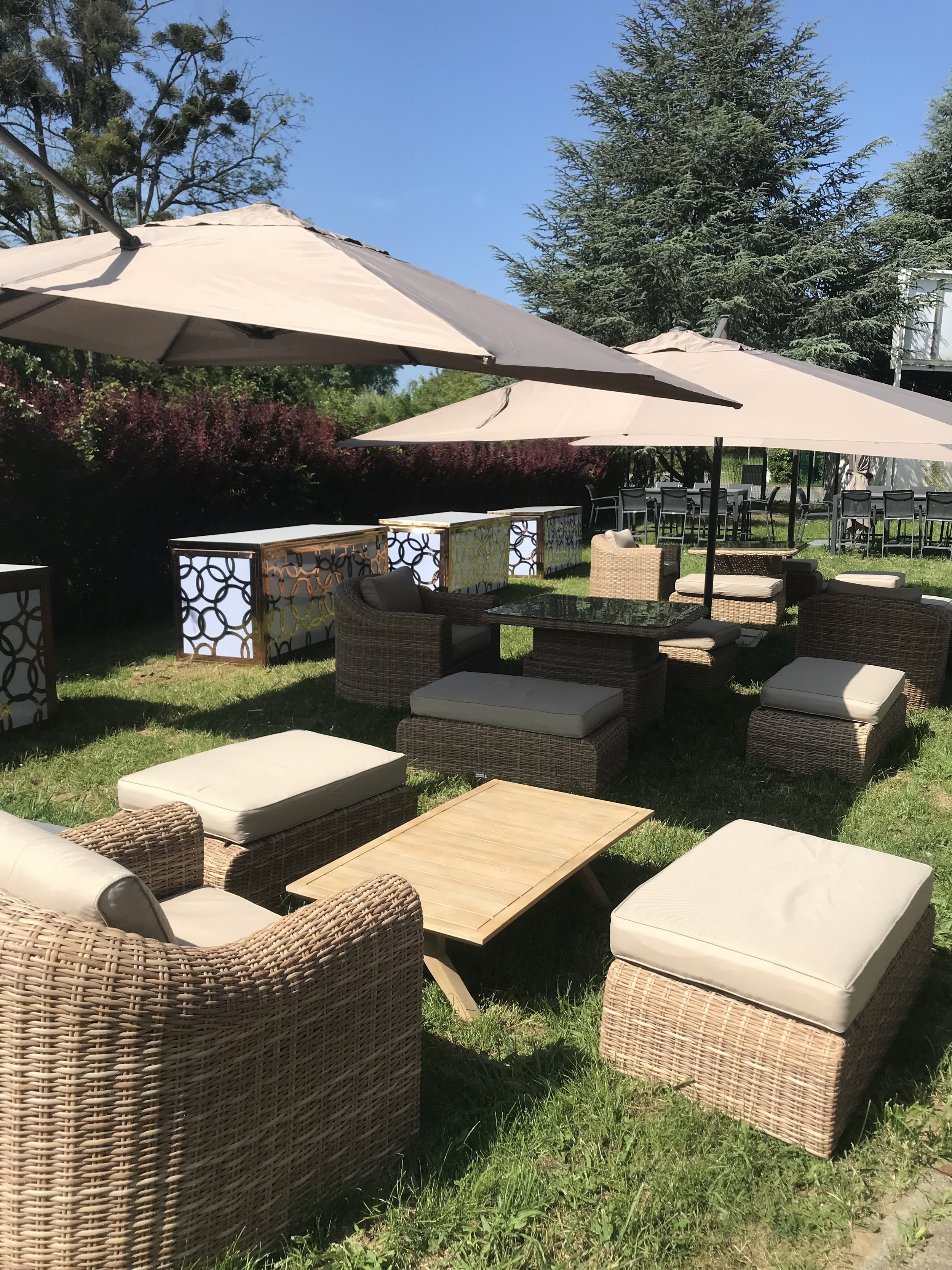Espace extérieur avec des fauteuils en rotin avec coussins, tables en bois, parasols beige, et verdure, probablement une terrasse de café ou restaurant