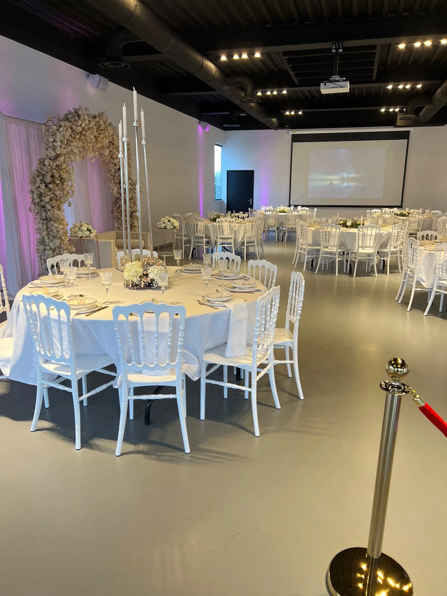Salle de réception décorée pour un mariage avec des tables rondes nappées de blanc, des chaises blanches, un centre de table floral et un grand écran.