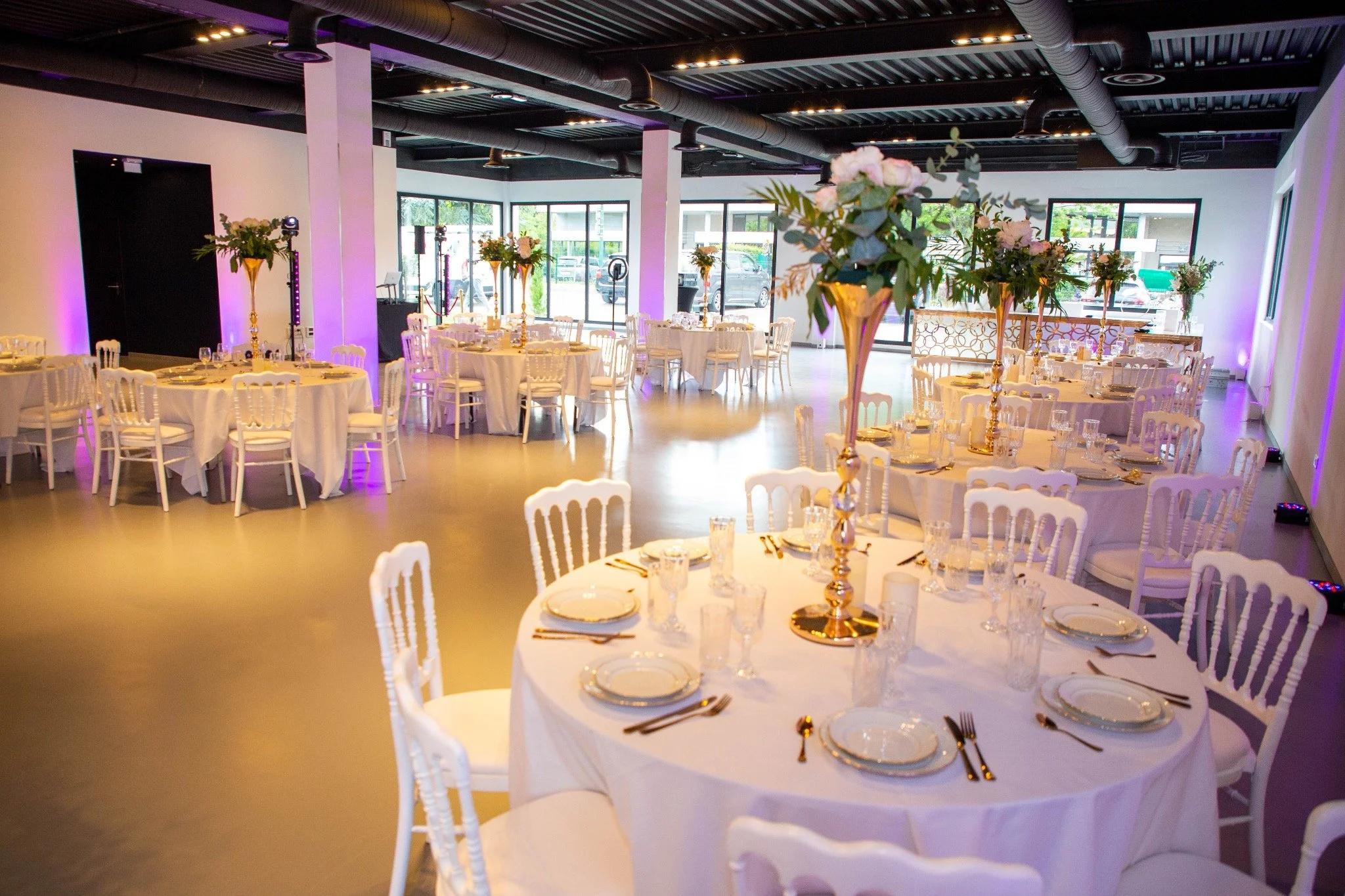 Salle de réception élégante avec des tables rondes habillées en blanc, décorées de hautes vases avec des arrangements floraux, dans un espace lumineux avec grandes fenêtres et un éclairage tamisé