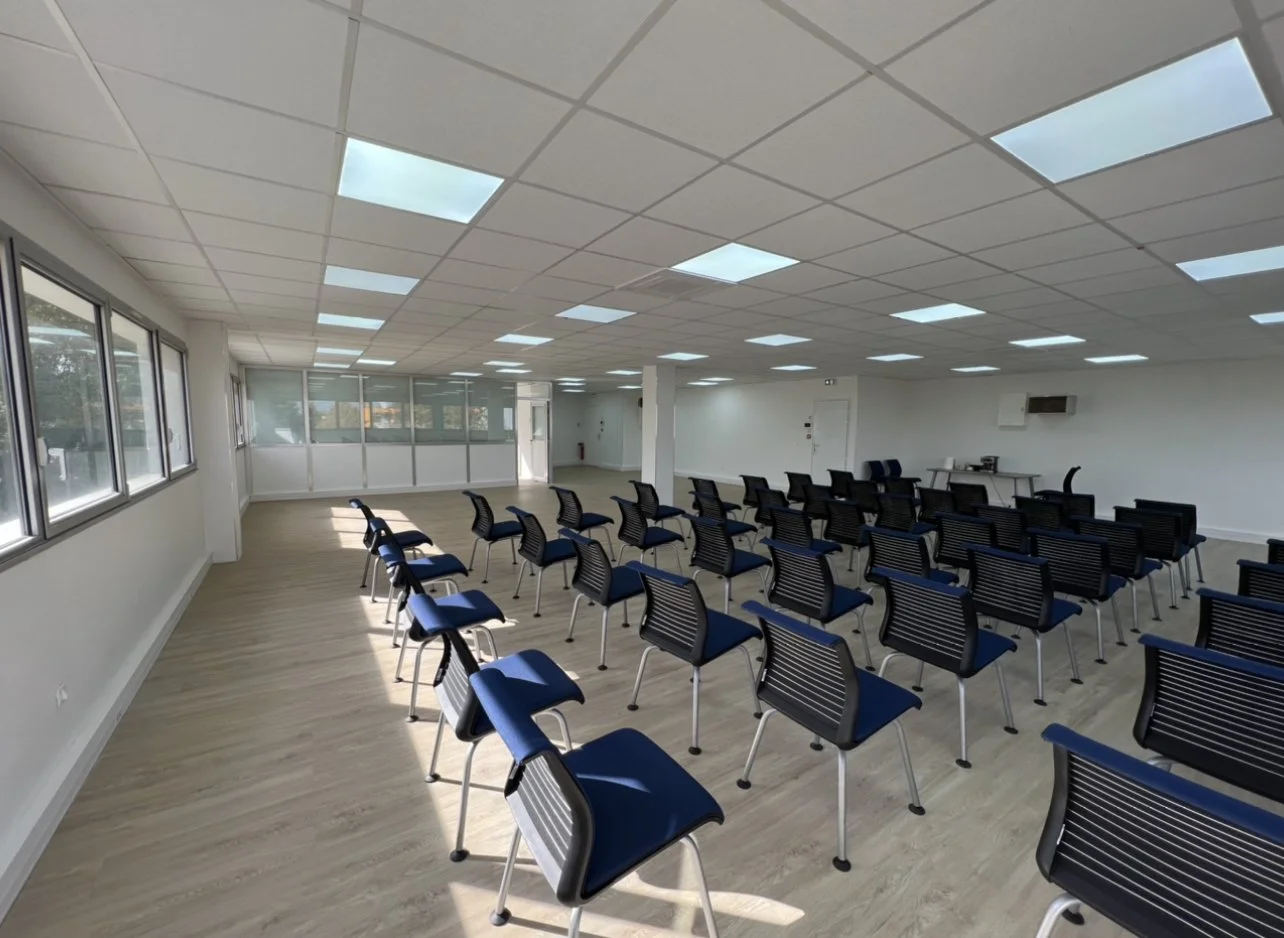 Salle de réunion vide équipée de plusieurs chaises noires avec coussin bleu, des fenêtres, un plafond avec des panneaux lumineux, et une table à l'arrière