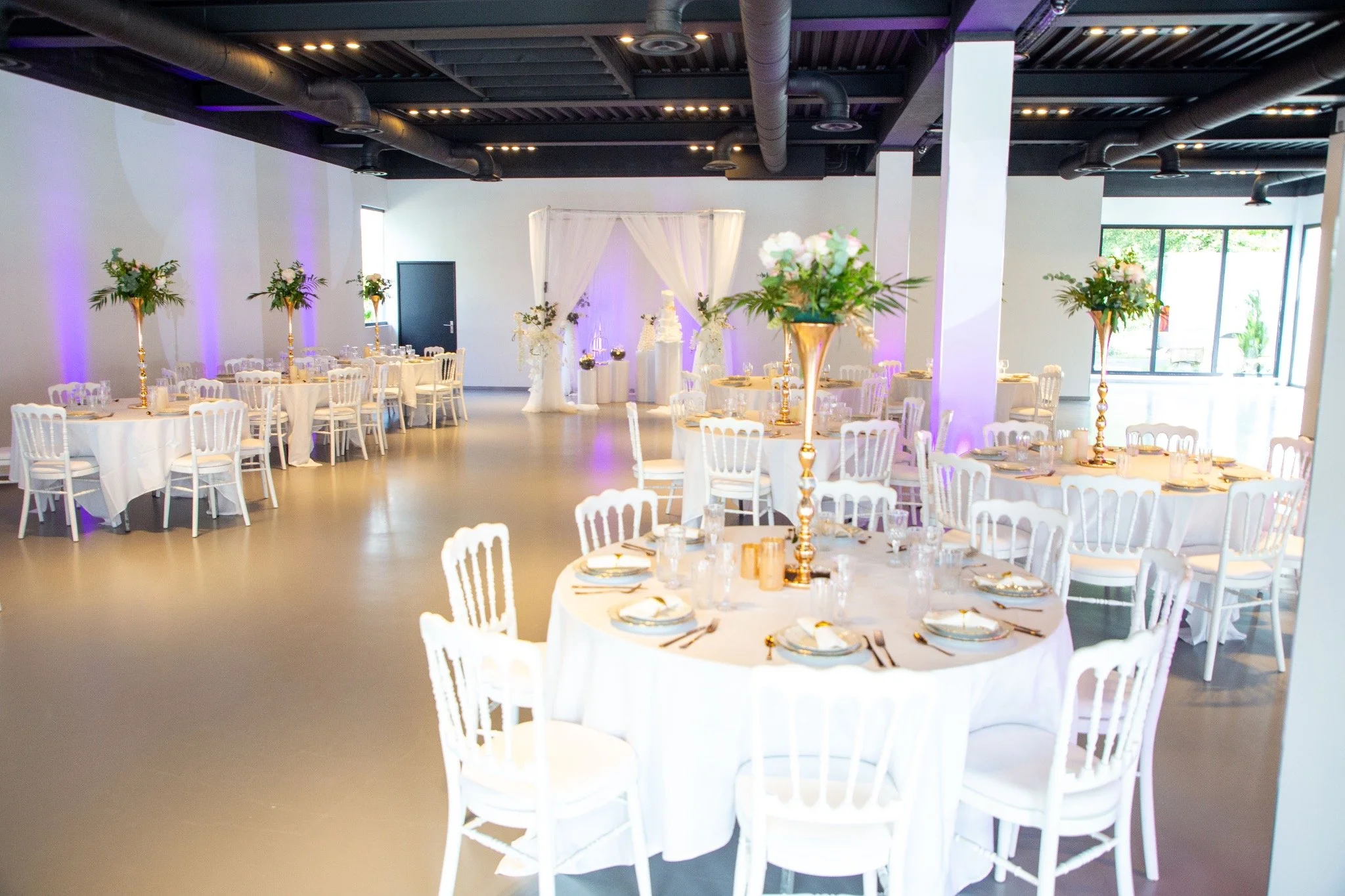 Salle de réception décorée pour un mariage avec des tables rondes ornées de nappes blanches et des centres de table en fleurs, avec des éclairages violets et une scène avec un cadre en drap blanc.