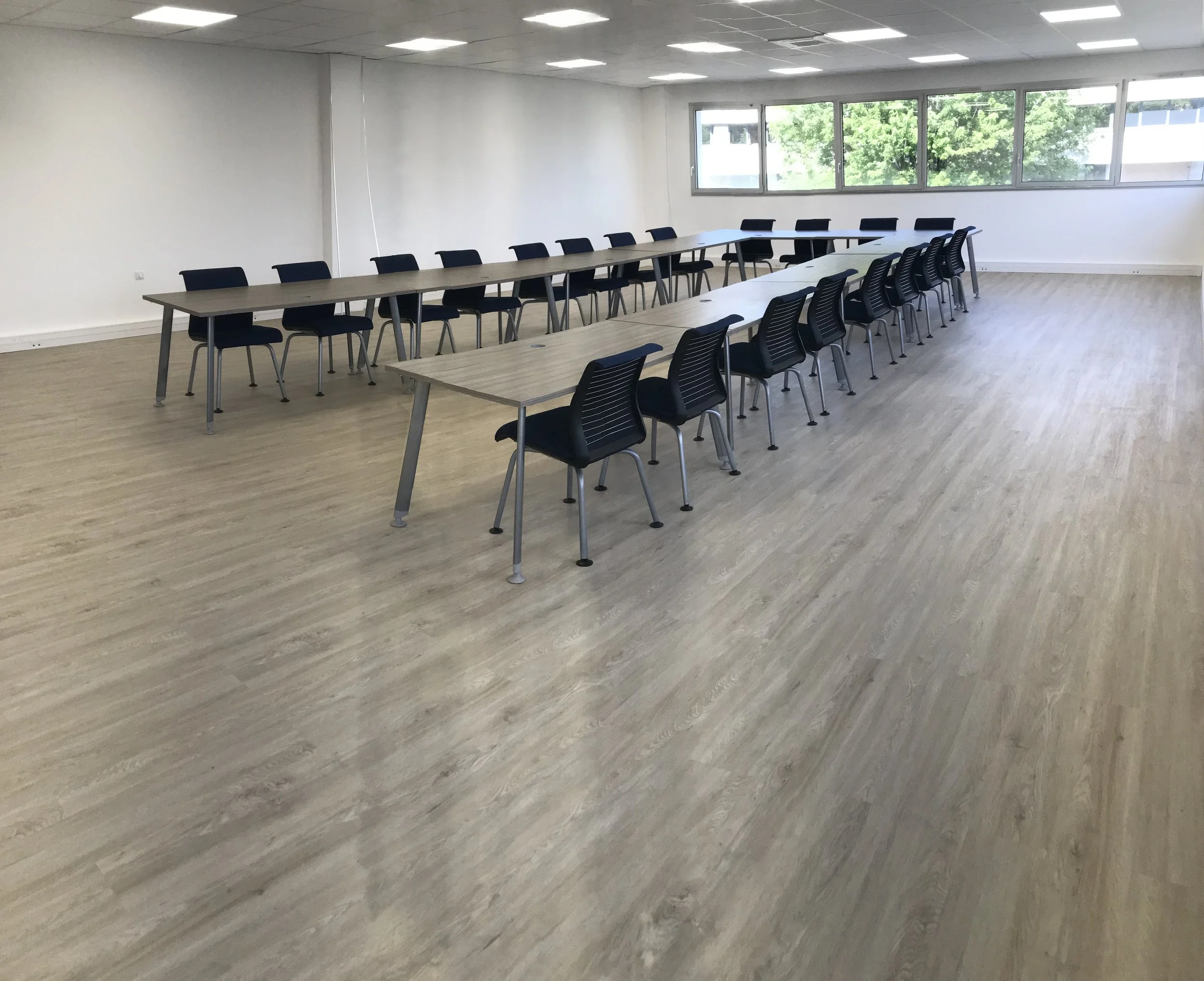Une grande salle de réunion avec des tables en bois et des chaises noires disposées en rangées, fenêtres laissant entrer beaucoup de lumière naturelle.