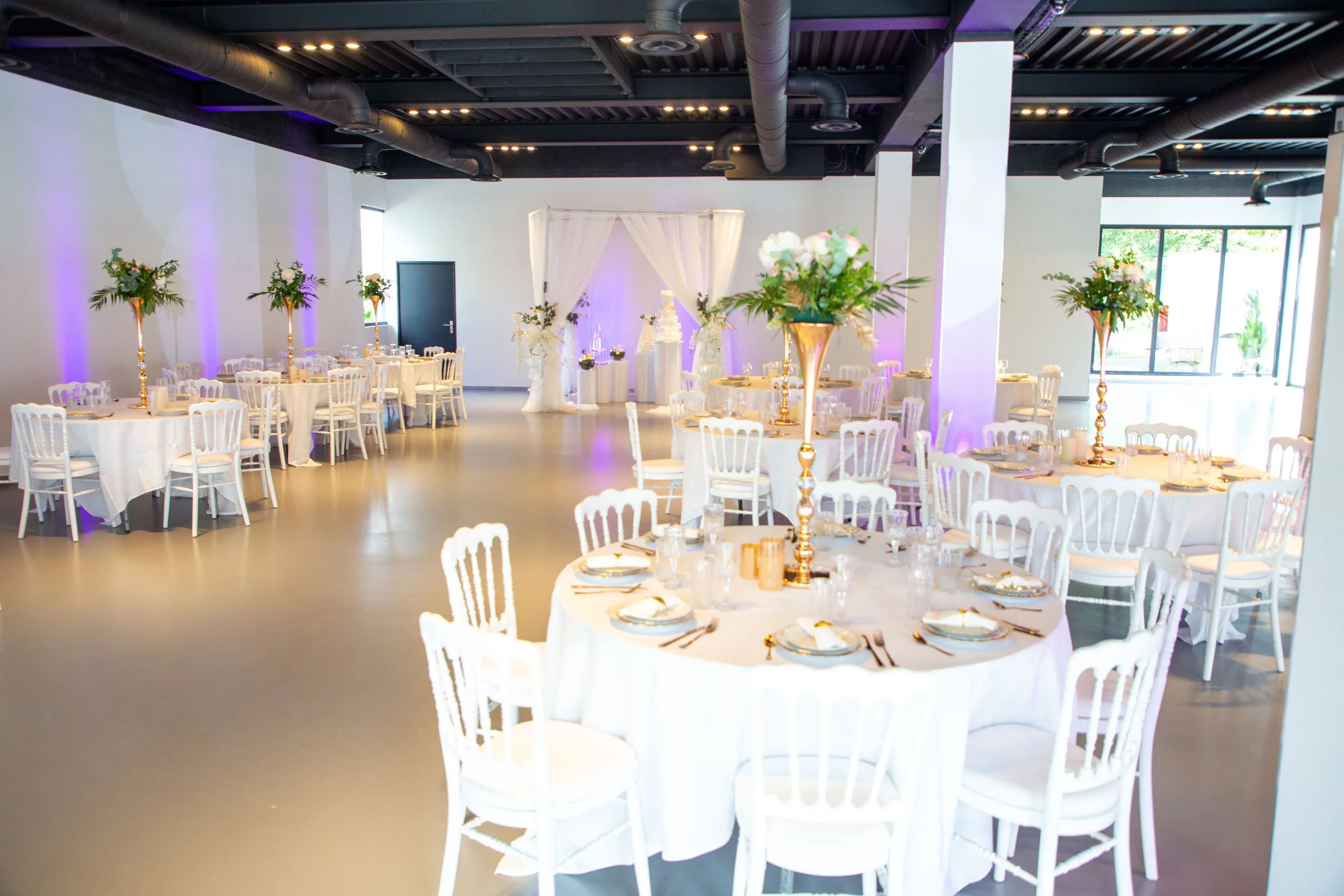Salle de réception décorée pour un mariage avec tables rondes, centres de table floraux, drapés blancs et éclairage violet
