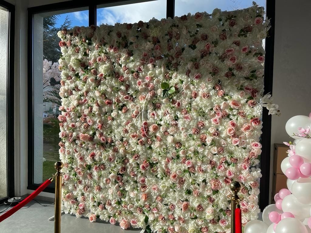 Un mur de fleurs composé principalement de roses blanches et roses, utilisé comme décoration pour un événement ou une occasion spéciale, situé près d'une grande fenêtre.