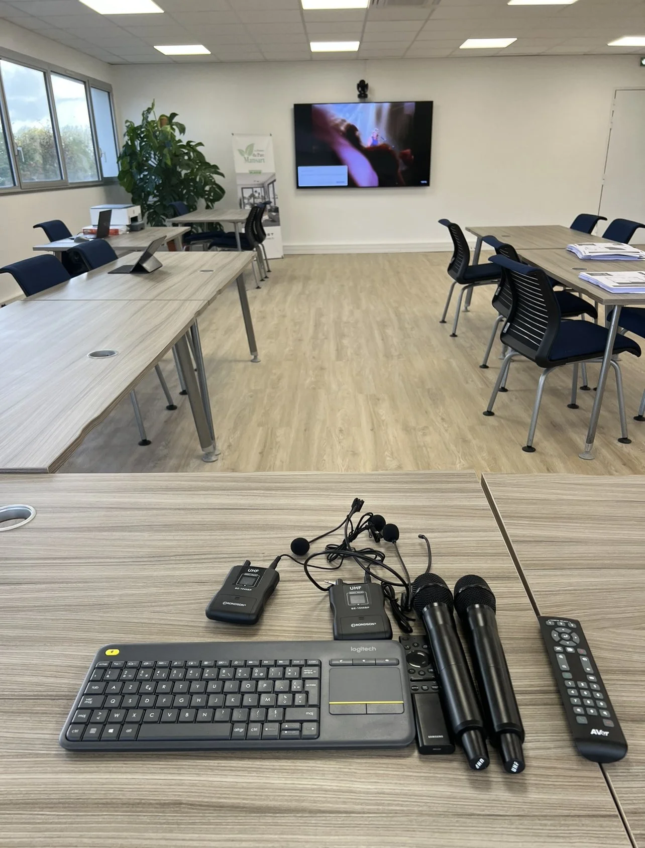 Salle de réunion avec tables, chaises, grand écran, et équipement audio dont microphones, télécommande, clavier, et microphones sans fil sur une table.