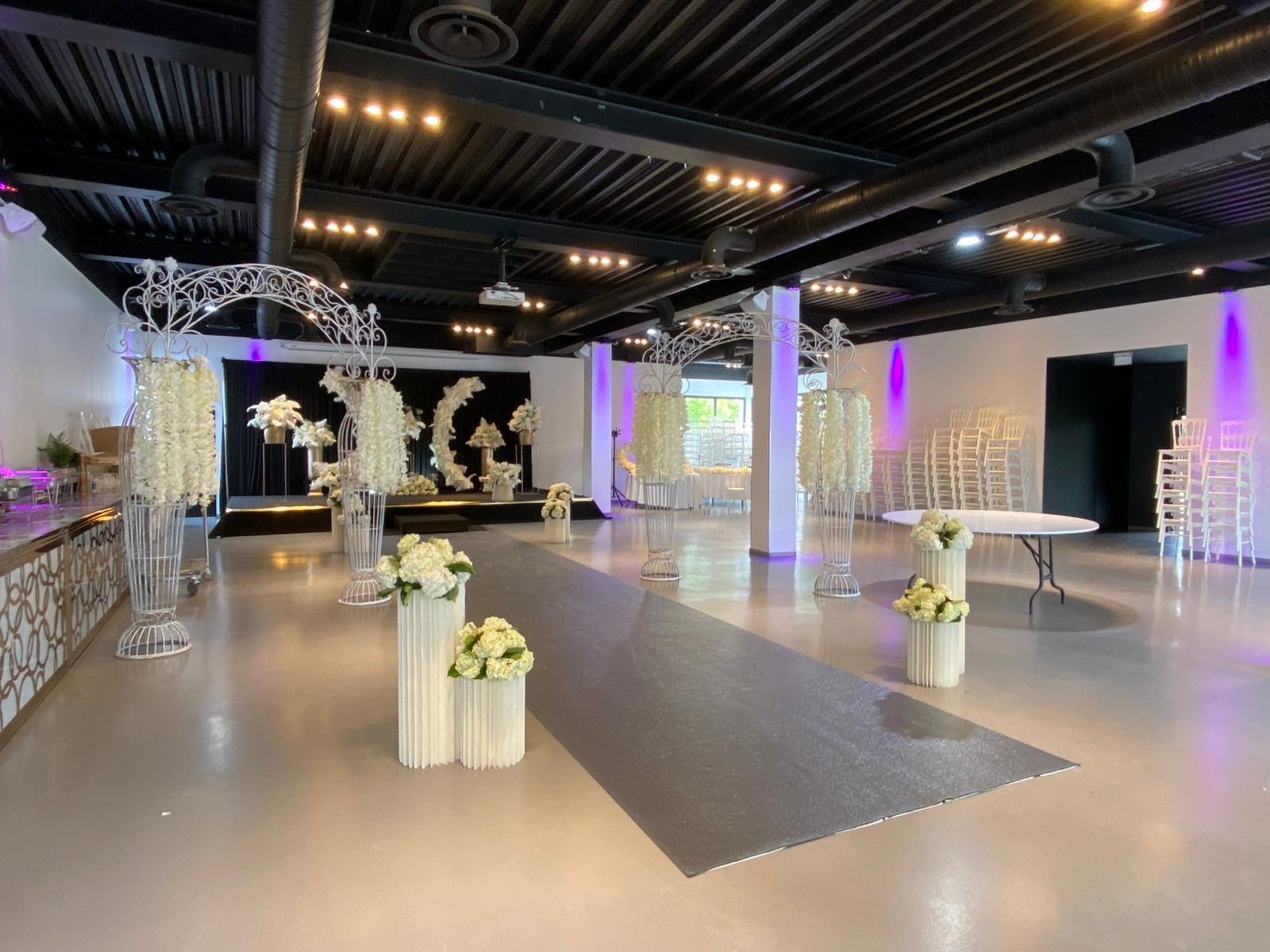 Salle de mariage décorée avec des arches en fer blanc ornées de fleurs blanches, plusieurs vases avec des bouquets de fleurs blanches, scène avec un fond noir pour la cérémonie, chaises empilées à droite, éclairage tamisé avec des lumières violettes.