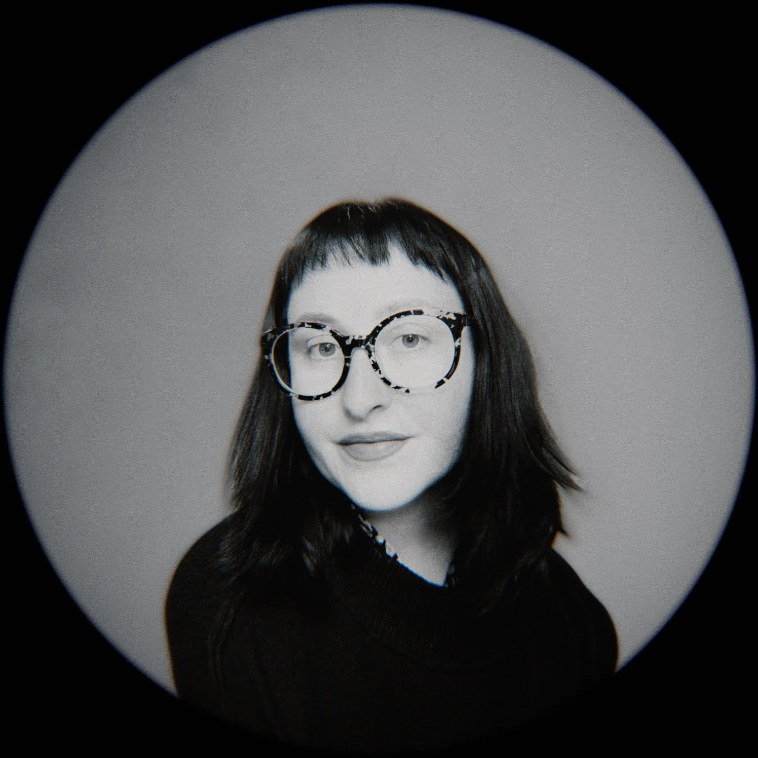 Black and white portrait of a woman with dark hair and large, round glasses, looking directly at the camera.