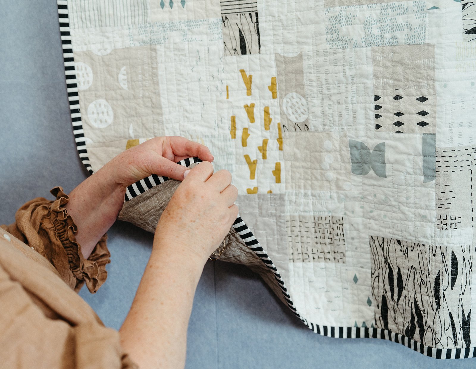Person sewing or quilting a patchwork quilt on a table, wearing a tan shirt and brown scrunchie on arm.