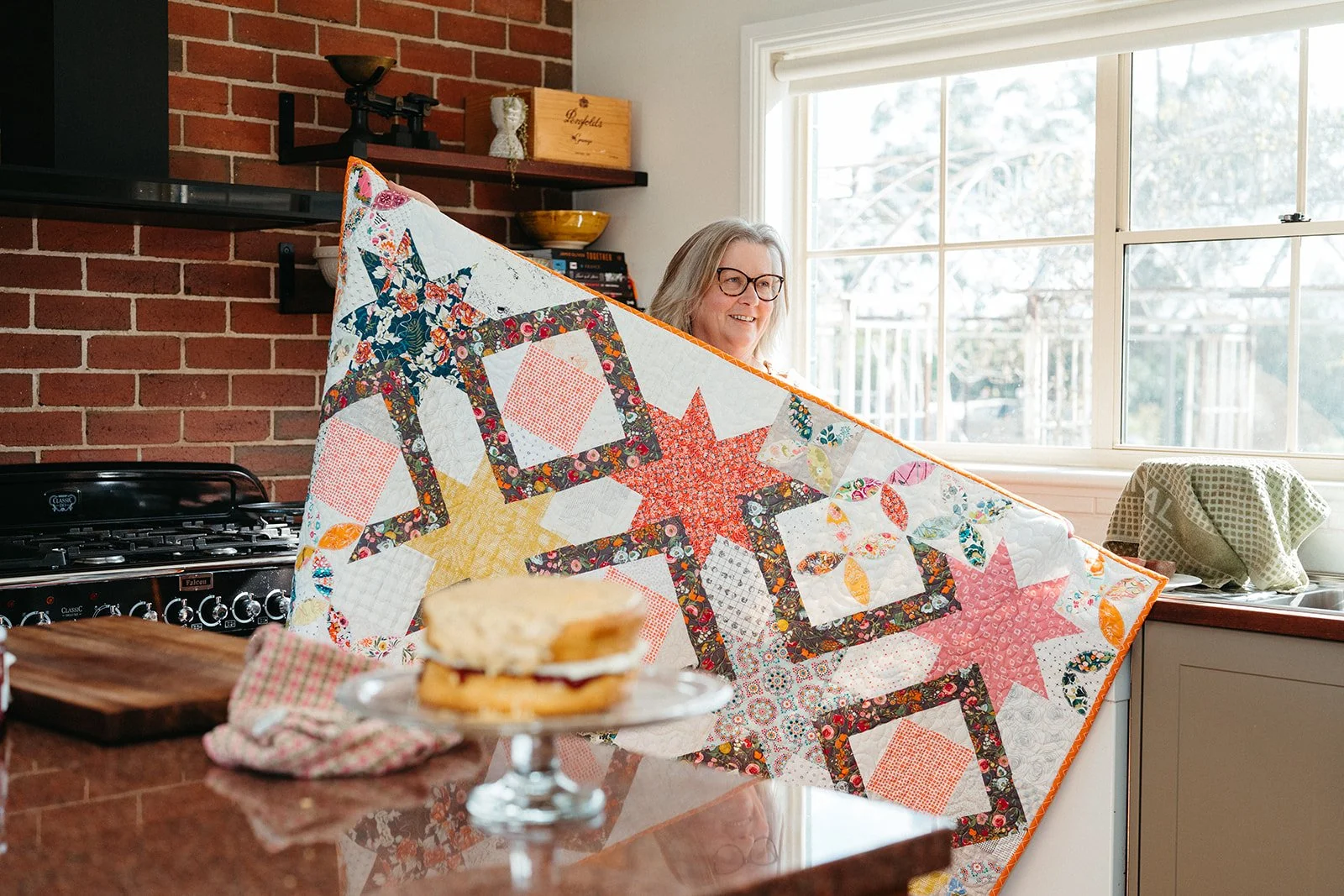 Karen holds up a custom quilt in her kitchen with her famous passionfruit sponge in the foreground.