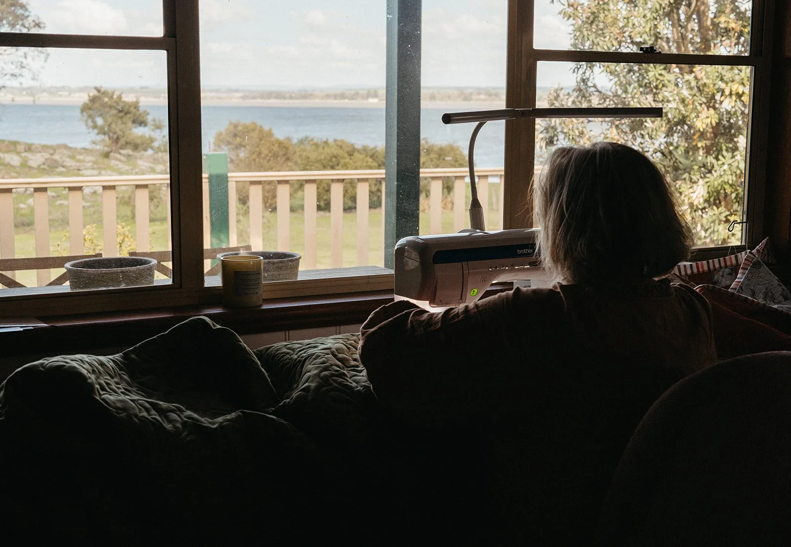 A woman sitting in a dark room, using a machine with a bright screen, facing a large window overlooking a body of water and trees, with some pots and a candle on the windowsill.