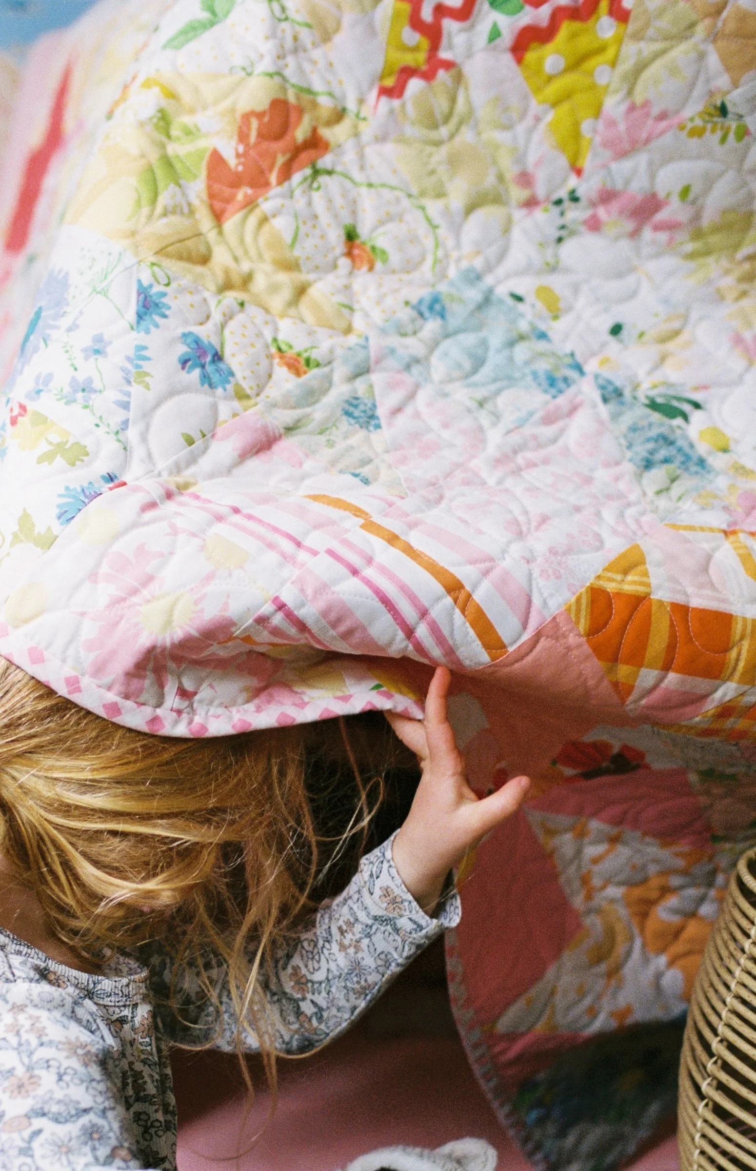 A young girl is reaching under a colorful quilt cubby with a floral pattern. The quilt features various pastel colors, including pink, blue, green, and yellow.