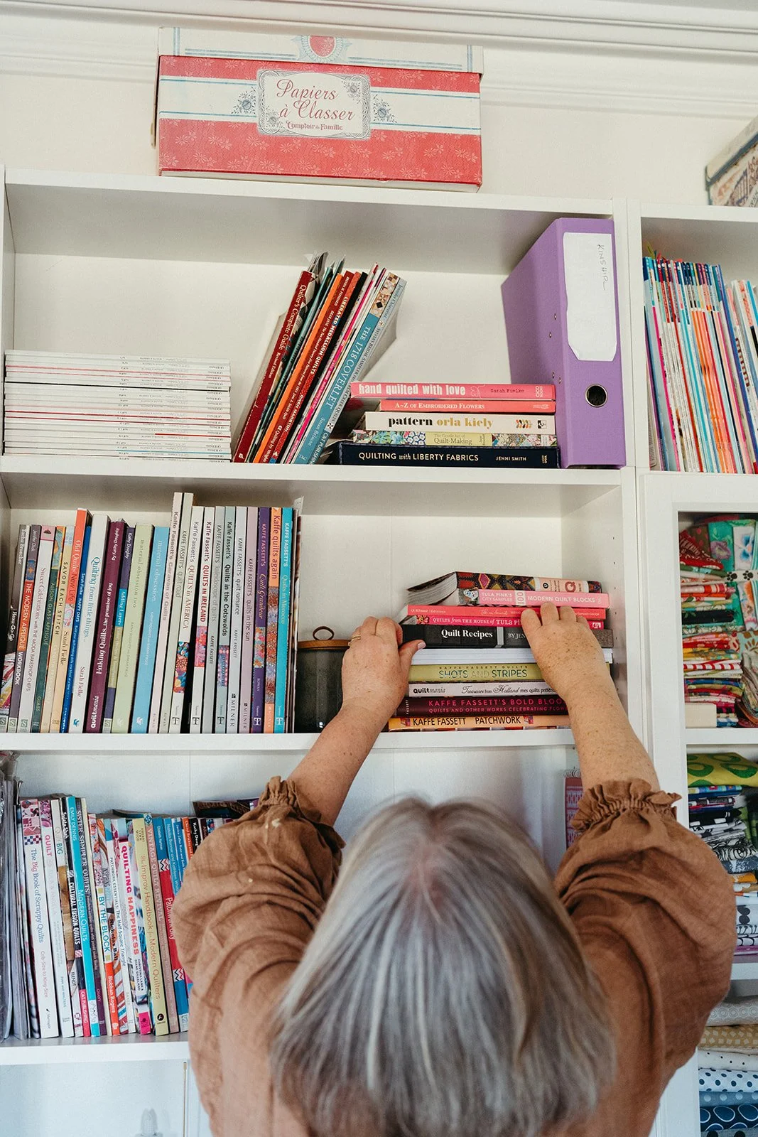 An elderly woman with gray hair, wearing a brown shirt, is reaching for a book on a white bookshelf filled with colorful books and craft supplies.