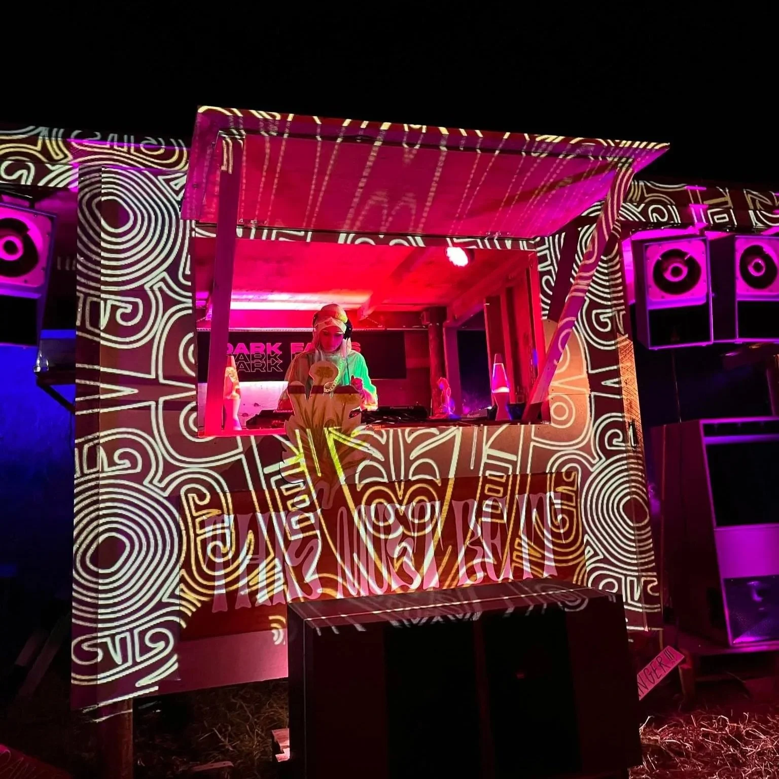 A DJ performing at This Must Be It in Raglan New Zealand with vibrant neon pink and purple lighting, and digital patterns projected onto the booth walls.