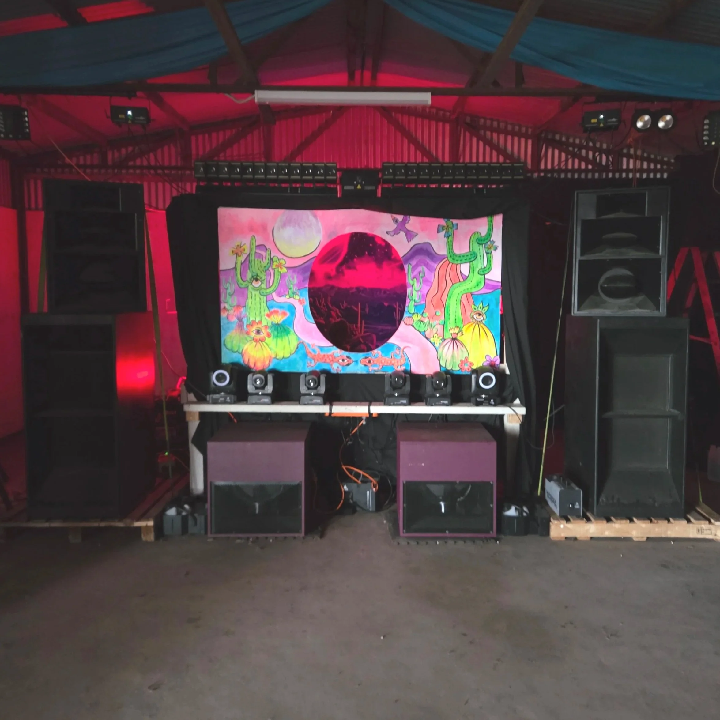 A stage setup with large black speakers on either side, a colorful digital backdrop featuring cacti, mountains, a full moon, and a circular image of a desert landscape with pink sky. The stage has various lighting equipment, and the floor is made of 
