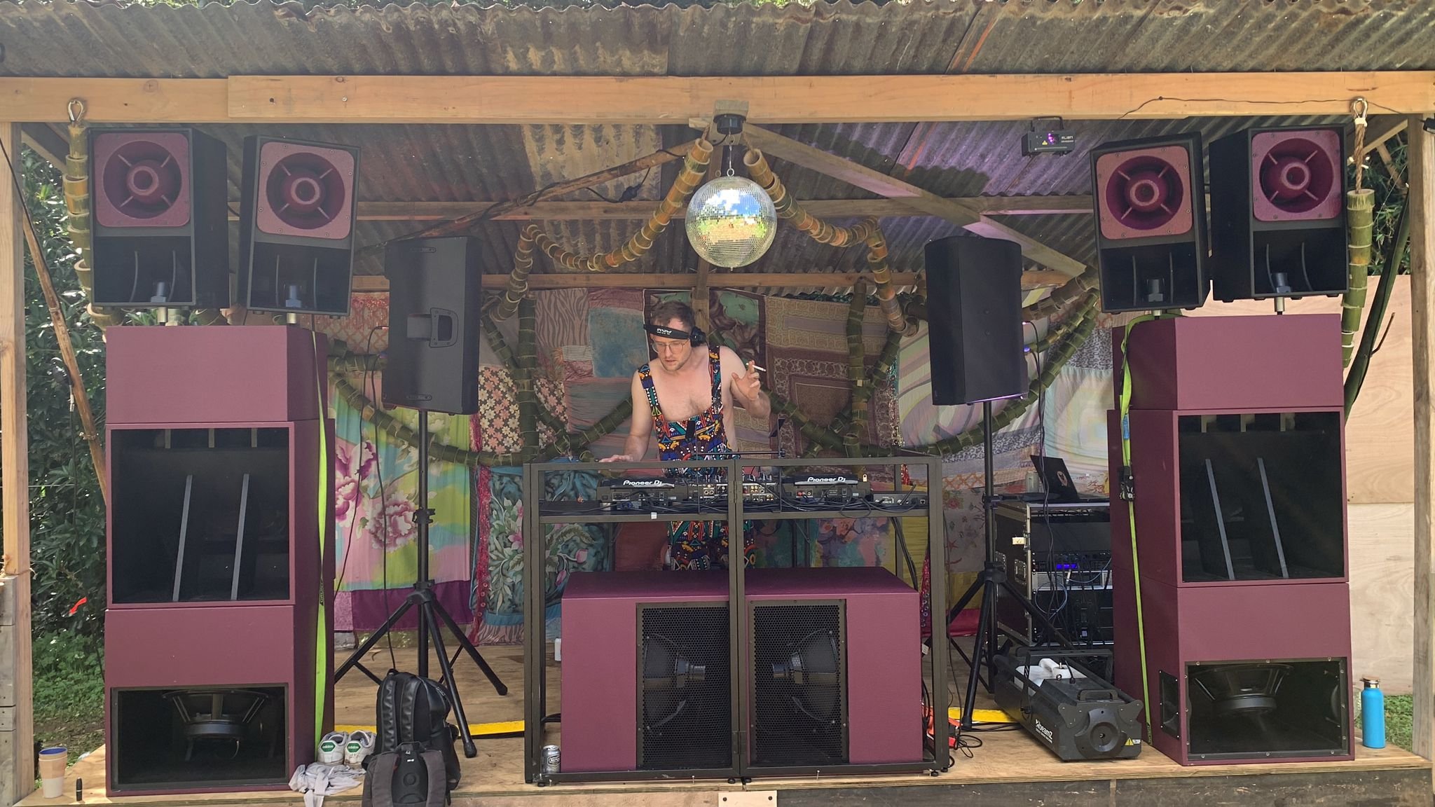 A DJ wearing colorful, patterned clothing and sunglasses performing on an outdoor stage with large pink and black speakers, a disco ball, and a backdrop of colorful fabric. The DJ is using equipment, with headphones on and one hand raised, in a rusti