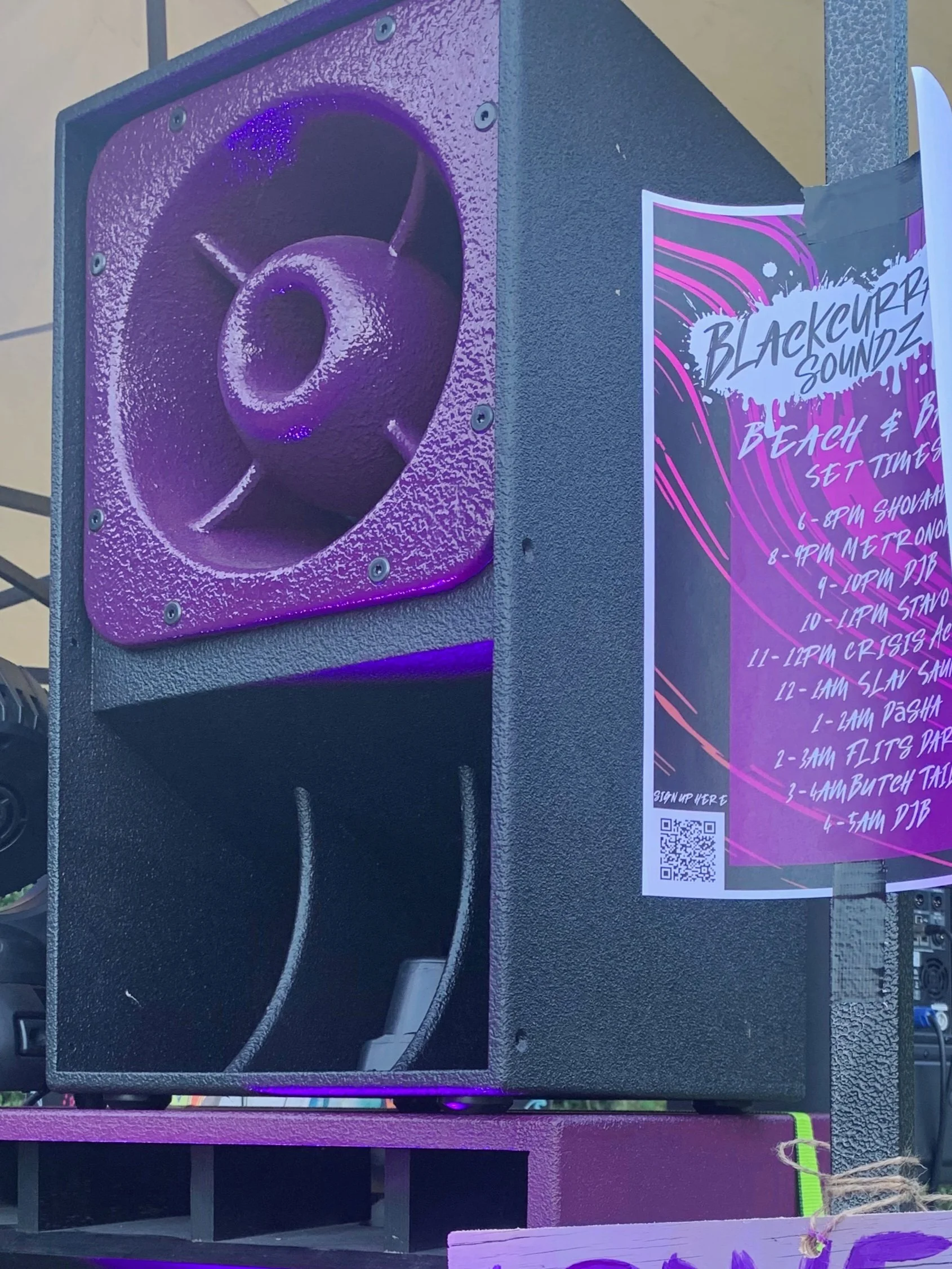 A large black speaker with a purple spinning horn on a purple stand. Next to it is a poster with a music event schedule titled 'Blackcurrant Soundz Beach & Bass' with set times listed.
