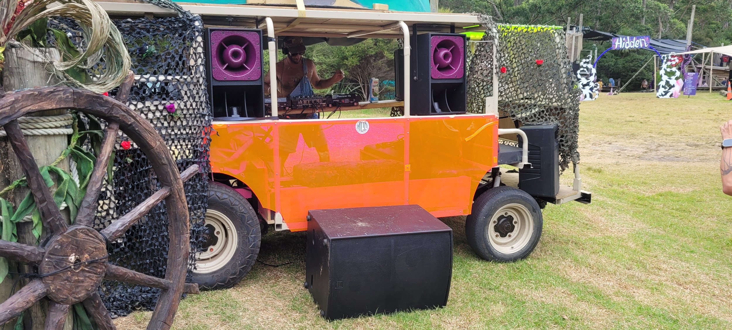 An outdoor music setup on a modified vehicle with DJ equipment, large purple speakers, and camouflage decorations, with a person DJing inside and an audience in the background.