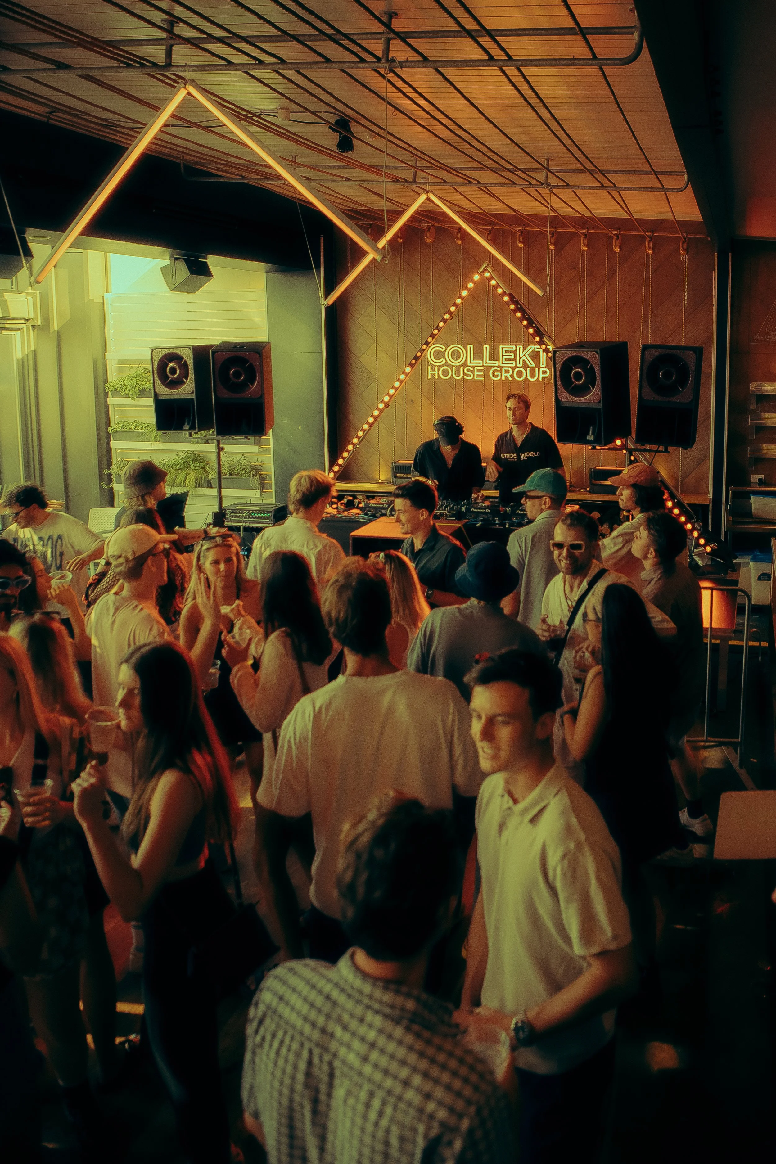 Crowd of people dancing and socializing at a DJ concert in an indoor venue with neon sign reading 'COLLEKT HOUSE GROUP' and modern lighting fixtures overhead.