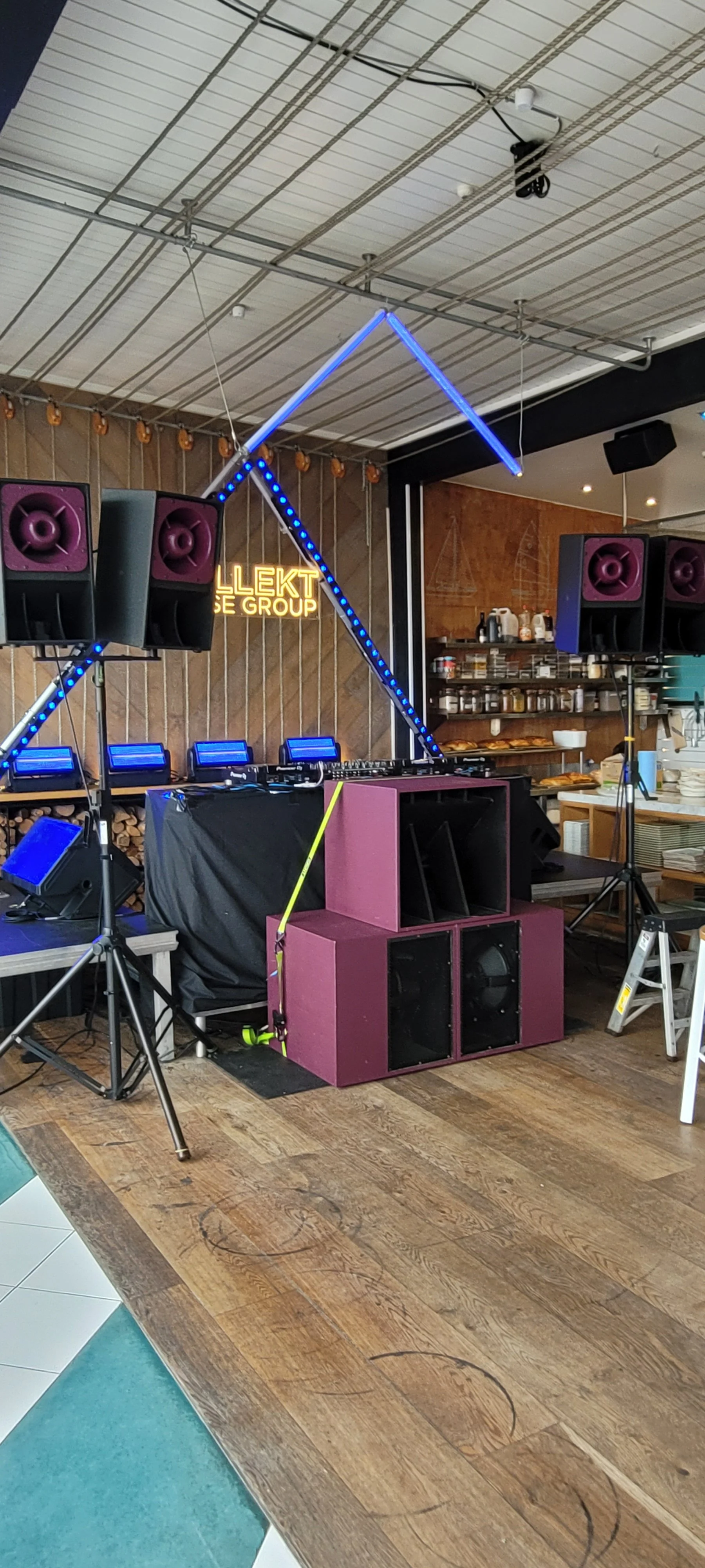 Music DJ setup with speakers, lighting, and a laptop on a table in a restaurant or event space with wood accents and shelves of food items in the background.