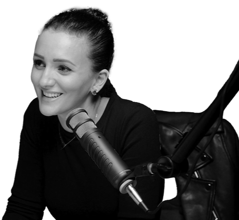 A woman with dark hair styled in a bun, smiling while speaking into a professional microphone, seated in a studio or radio station setting.