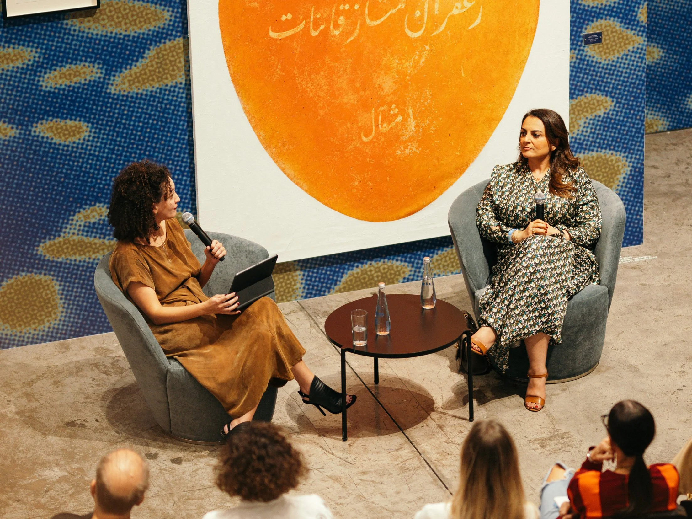 Two women seated in chairs on stage, engaged in conversation during a panel discussion at an event. One woman has curly hair and is dressed in a brown outfit, holding a microphone and a tablet. The other woman has long curly hair, wearing a patterned