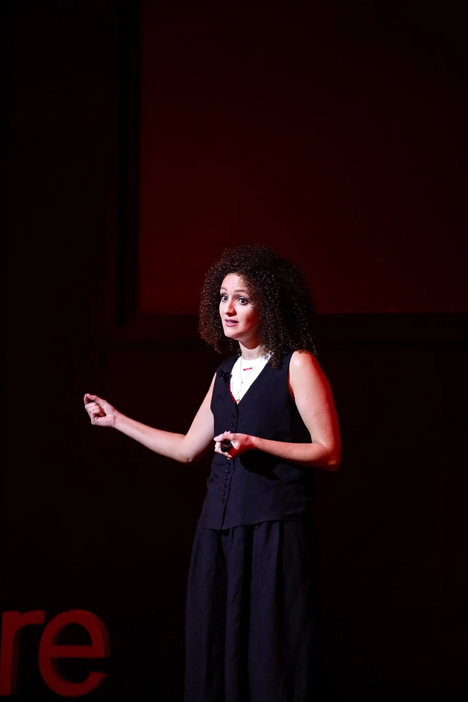 A woman with curly hair giving a presentation on stage, wearing a sleeveless black top and skirt, with a red background.