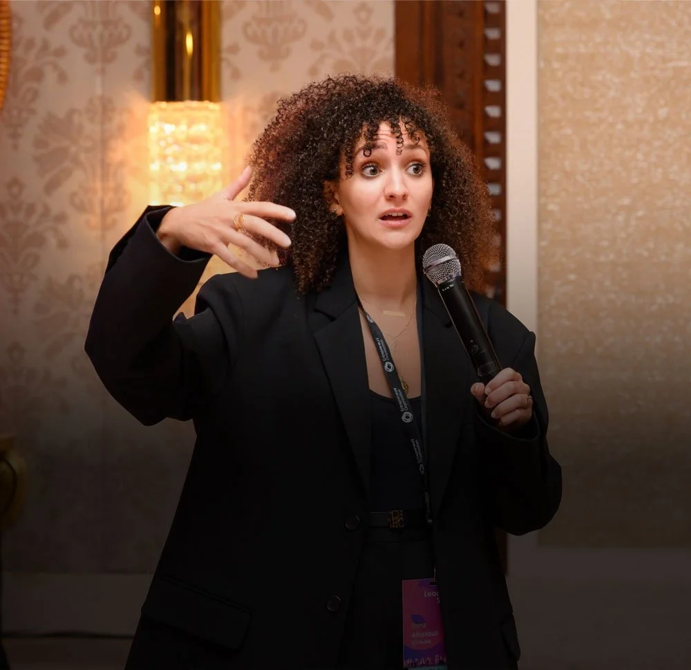 A woman with curly hair wearing a black blazer, speaking into a microphone, gesturing with her hand during a presentation or speech.