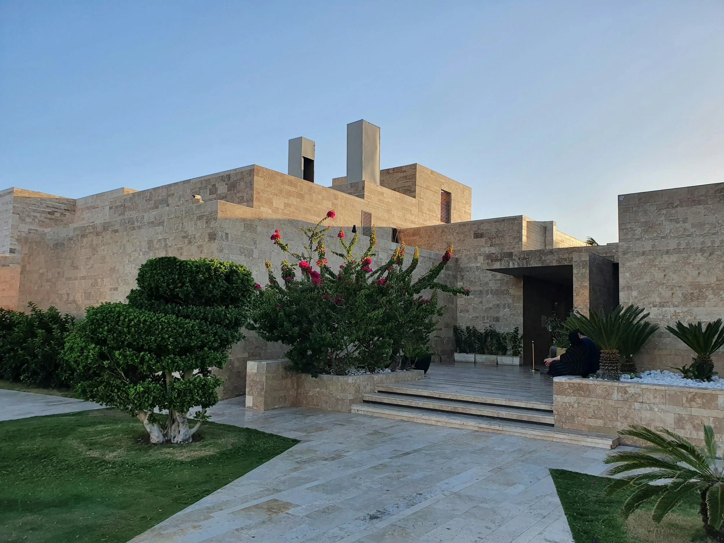 Modern beige stone building with steps, greenery, and a person sitting outside at sunset.