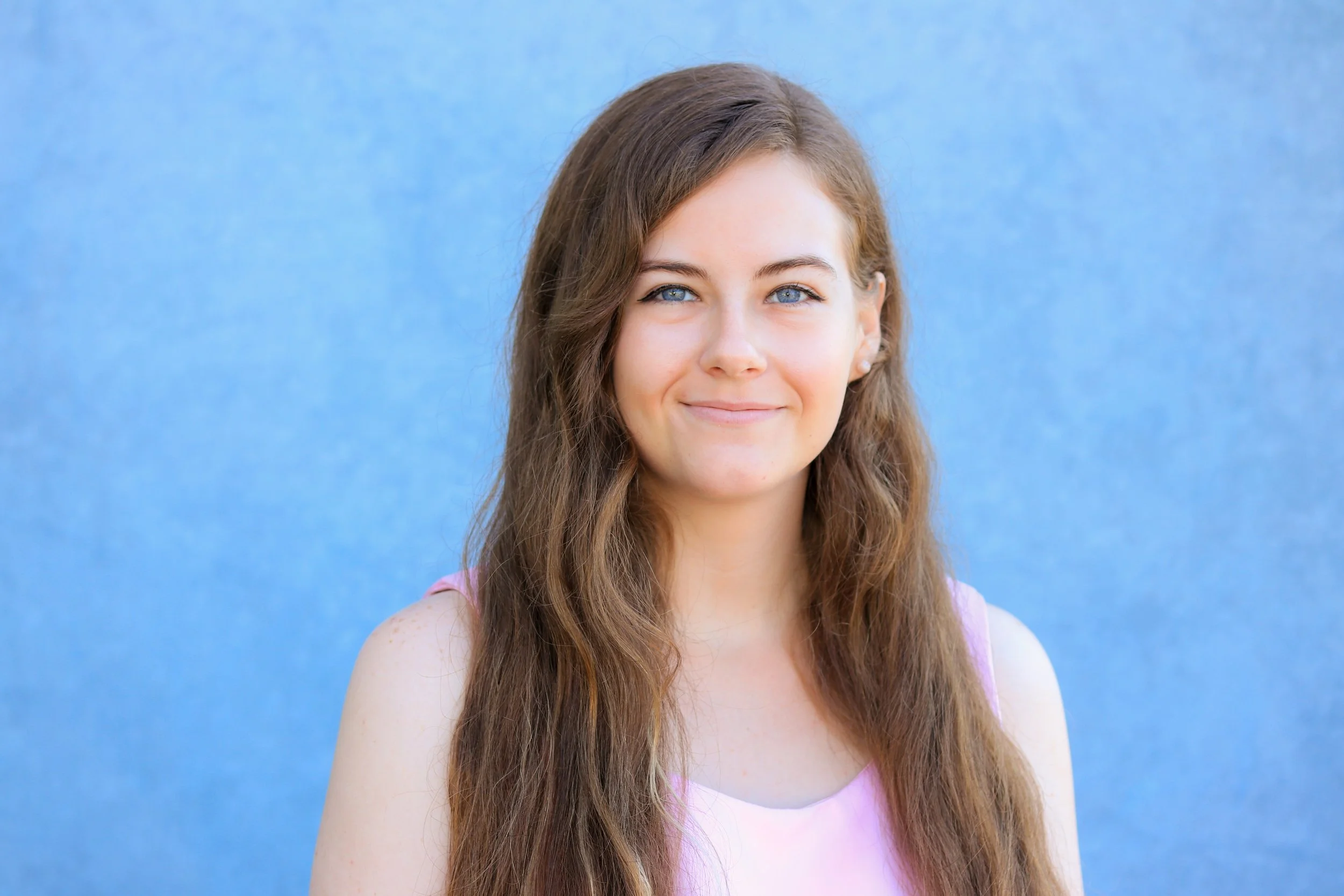Portrait of a young woman with long, wavy brown hair, blue eyes, and a light pink sleeveless top, standing against a blue background.