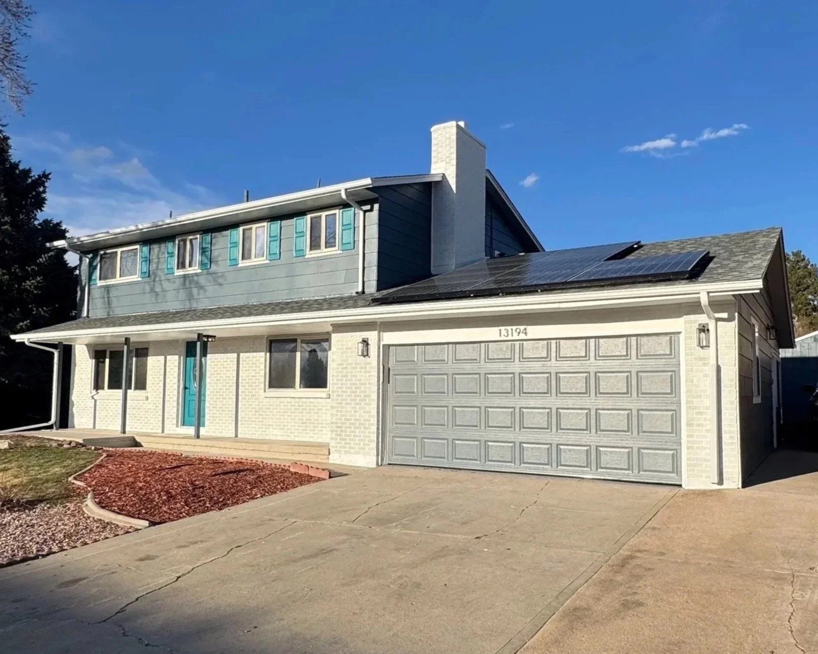 A two-story house with a light-colored brick exterior, teal front door, attached garage, and solar panels on the roof under a clear blue sky.