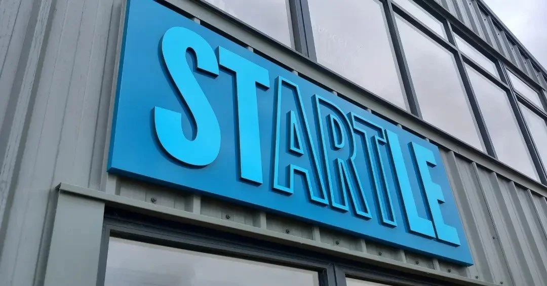 Blue sign says 'STARTLINE' in front of a modern building with large windows and gray metal siding.