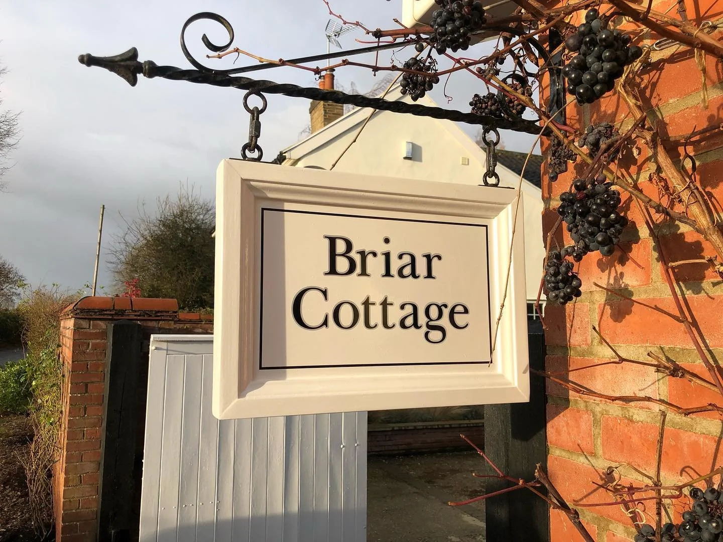 The image shows a hanging sign that reads 'Briar Cottage' in black lettering on a white background, attached to a black iron bracket over a red brick wall. There are grapevine and dark grapes growing on the brick wall on the right side, and trees and buildings in the background under a cloudy sky.