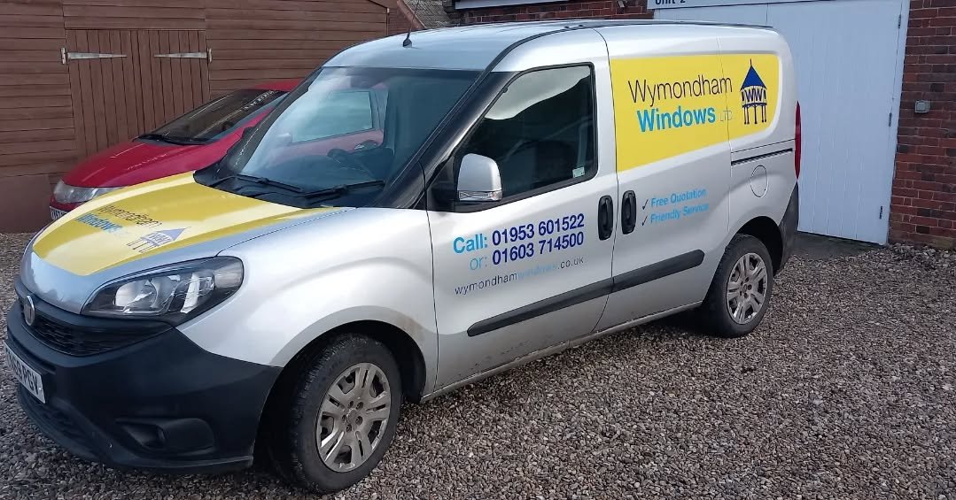 A silver van with Wymondham Windows Ltd logo and contact information parked on a gravel driveway.