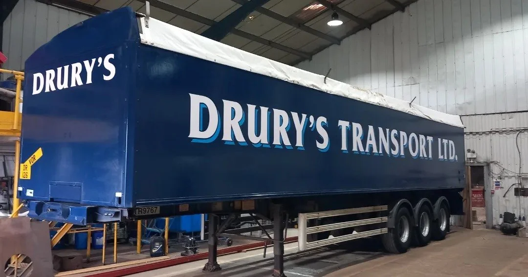 A large blue semi-trailer with white text reading DRURY'S TRANSPORT LTD. inside a warehouse or garage, with various equipment and support structures visible around it.