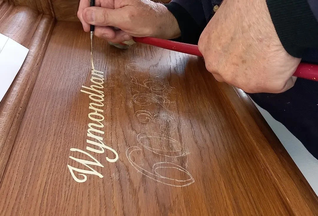 Person using a fine brush to paint the name 'Wymondham' onto a wooden surface as part of signwriting work.