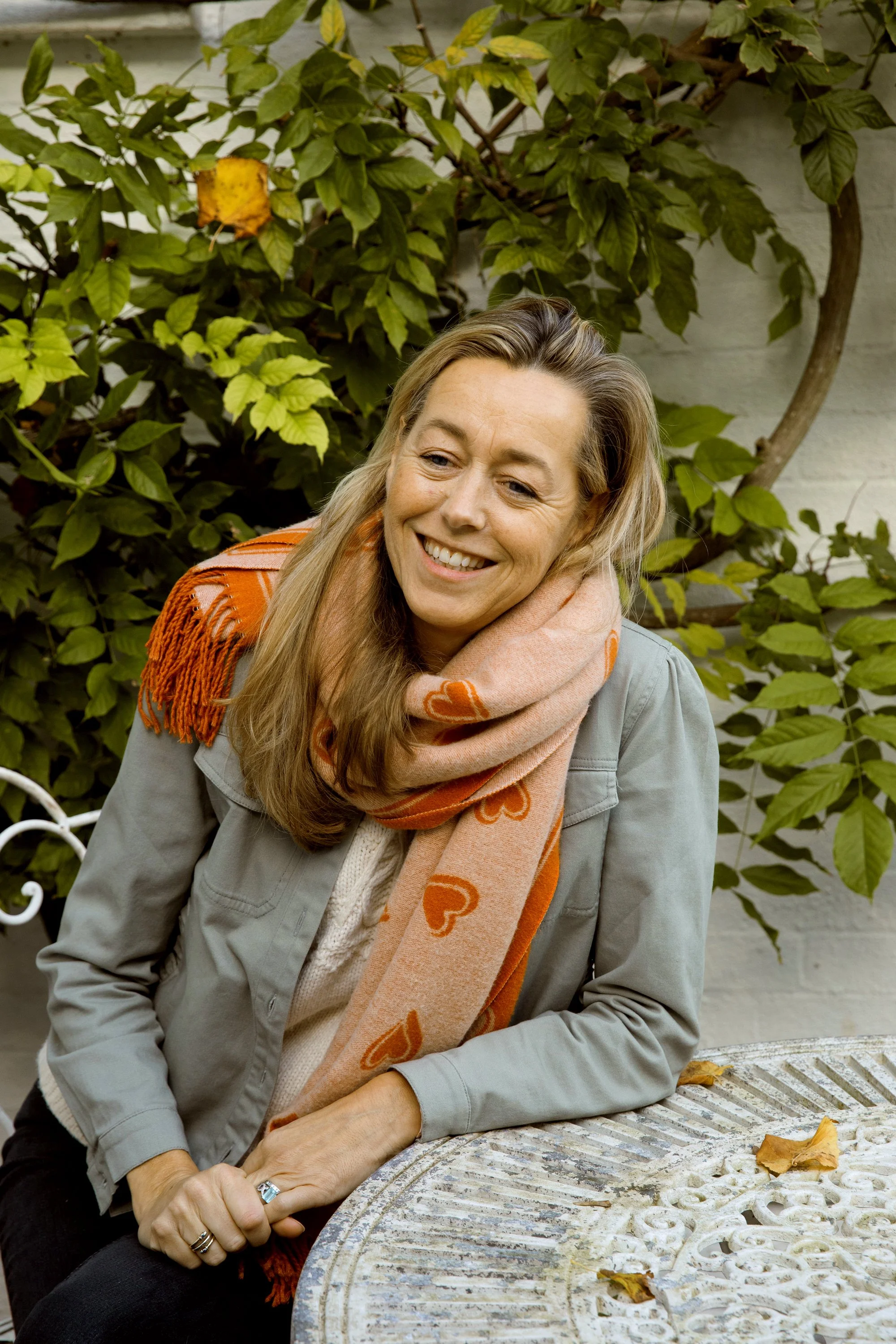 A woman with long, wavy blonde hair, wearing a gray jacket and a peach-colored scarf with orange hearts, smiling and sitting at a weathered metal table outdoors, surrounded by green leaves and a white brick wall.