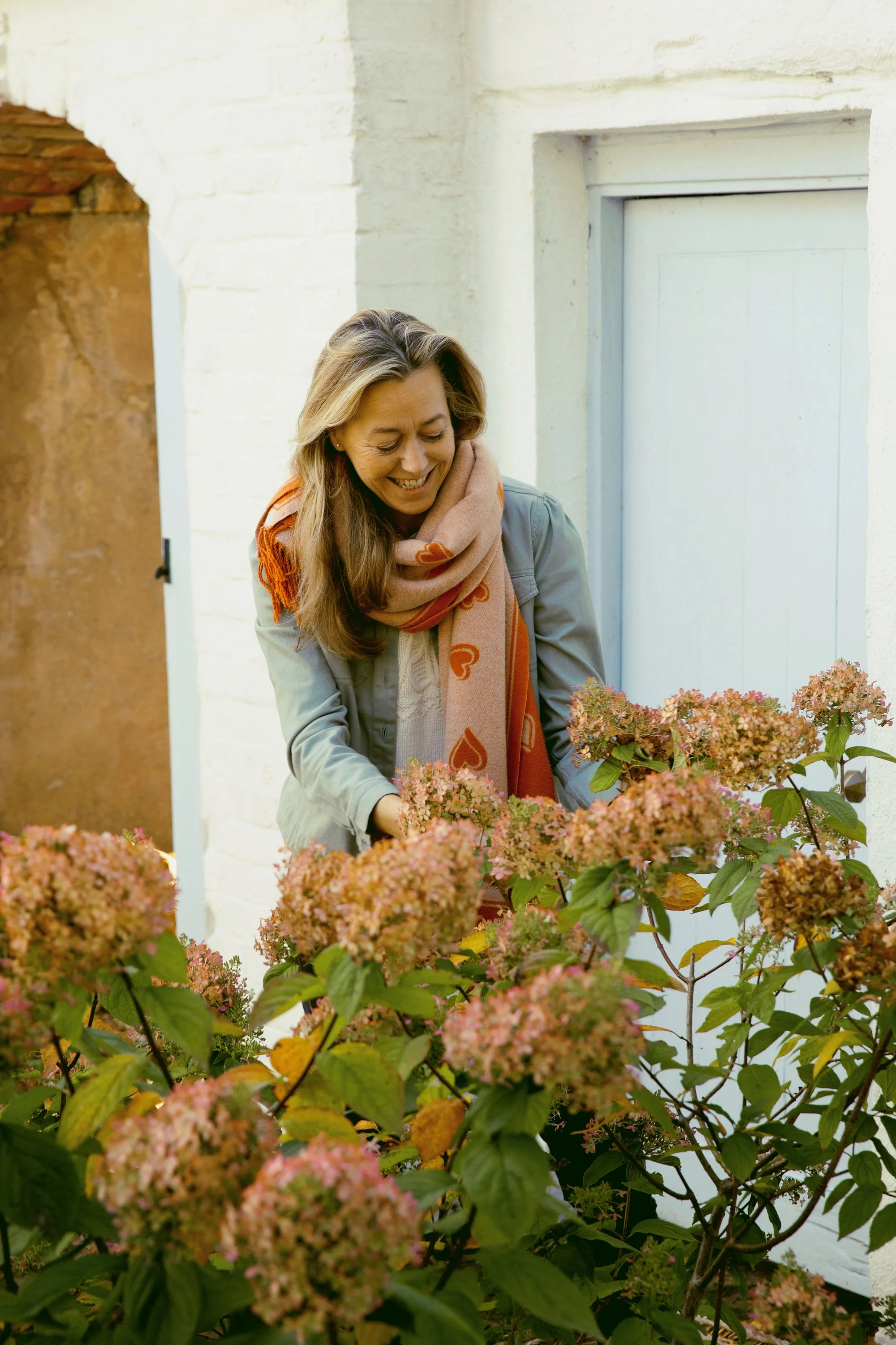 A woman in a light-colored coat and a pink scarf with orange hearts is smiling as she tends to pink and green hydrangea flowers outside a white-painted building with a window.