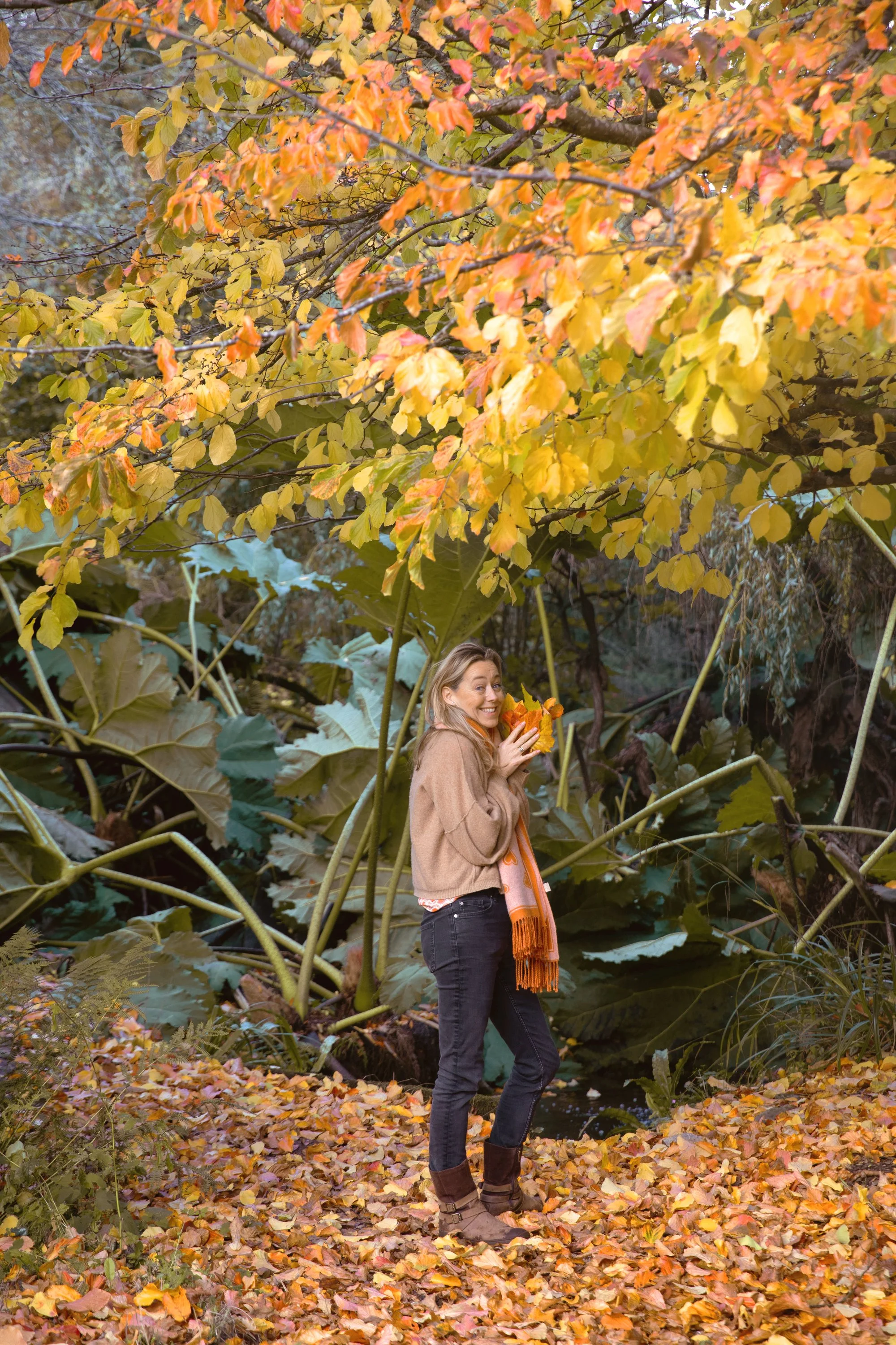 A woman in a tan jacket and black jeans standing on a leaf-covered path in a forest, holding autumn leaves and smiling.