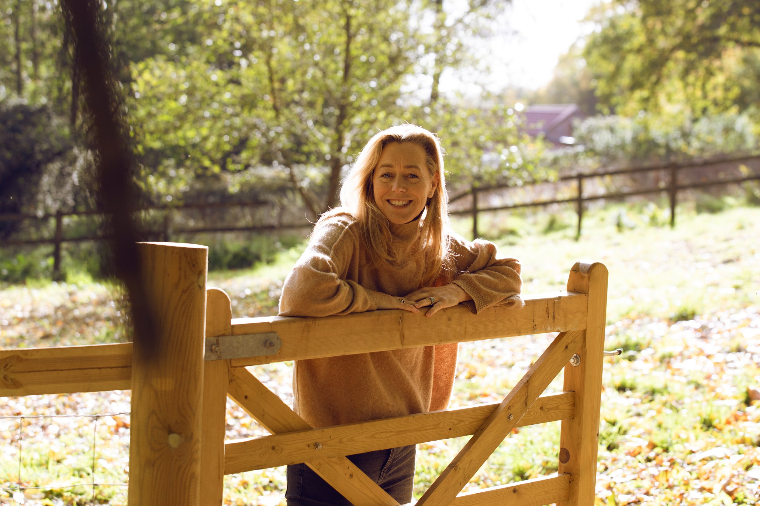 A woman with blonde hair smiling, leaning on a wooden gate outdoors with green trees and sunlight in the background.