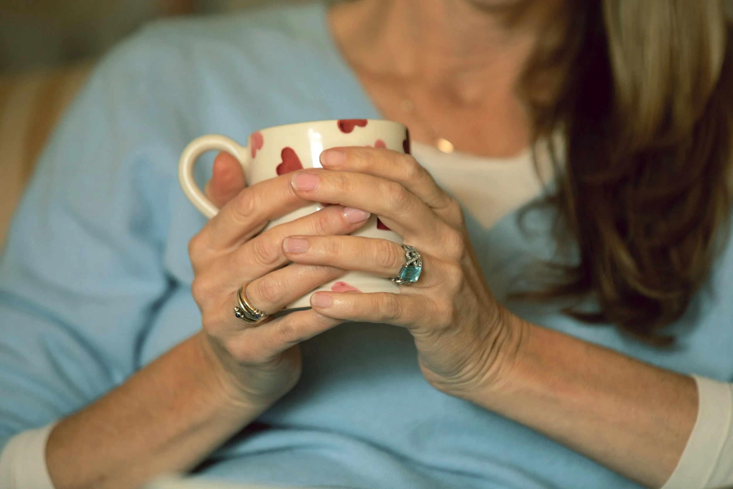 A woman with long brown hair holding a white mug with red hearts, wearing rings on her fingers, dressed in a light blue top.