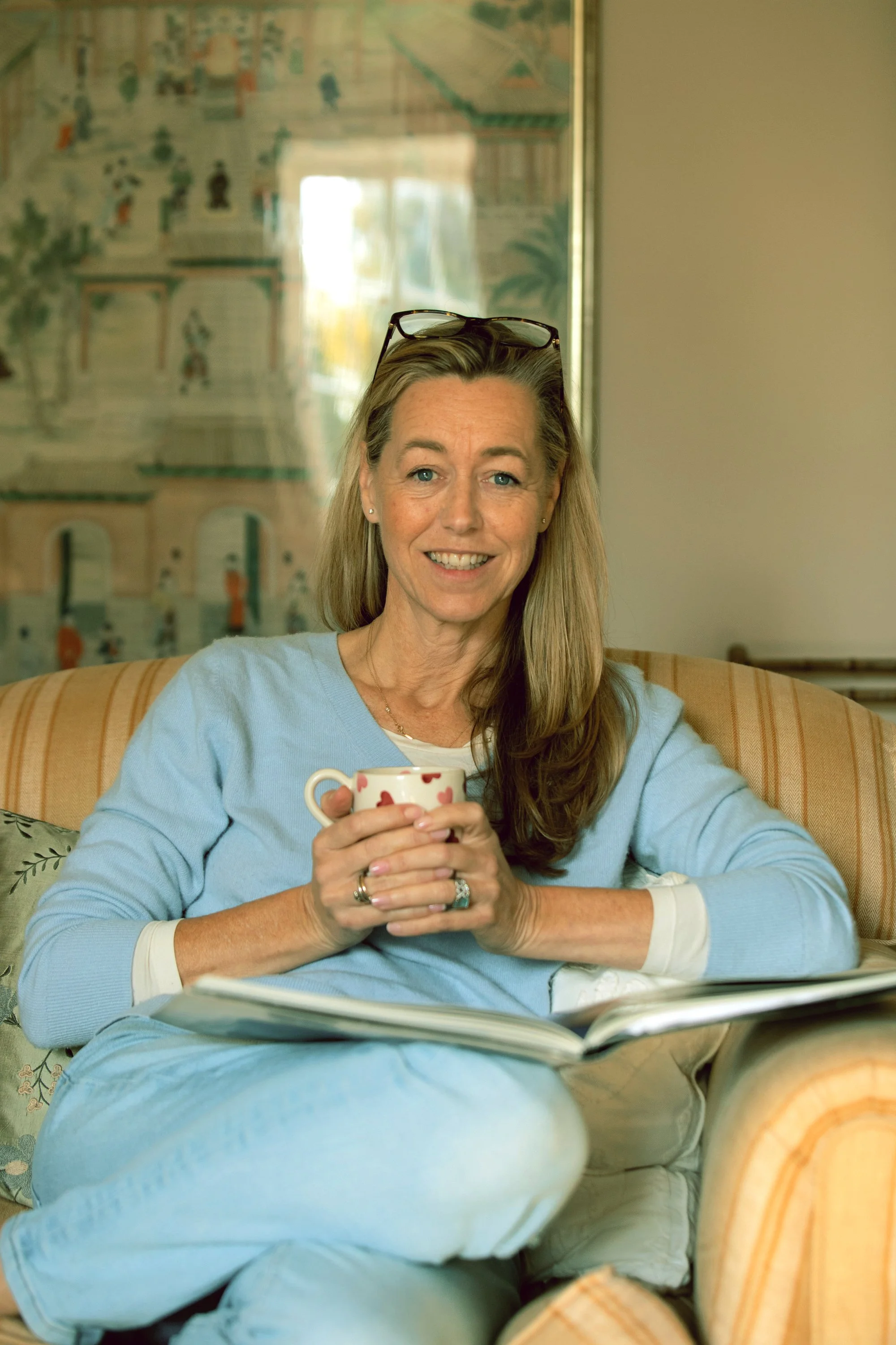 A woman with glasses on her head sitting on a couch, smiling, holding a cup, with an open book resting on her lap.