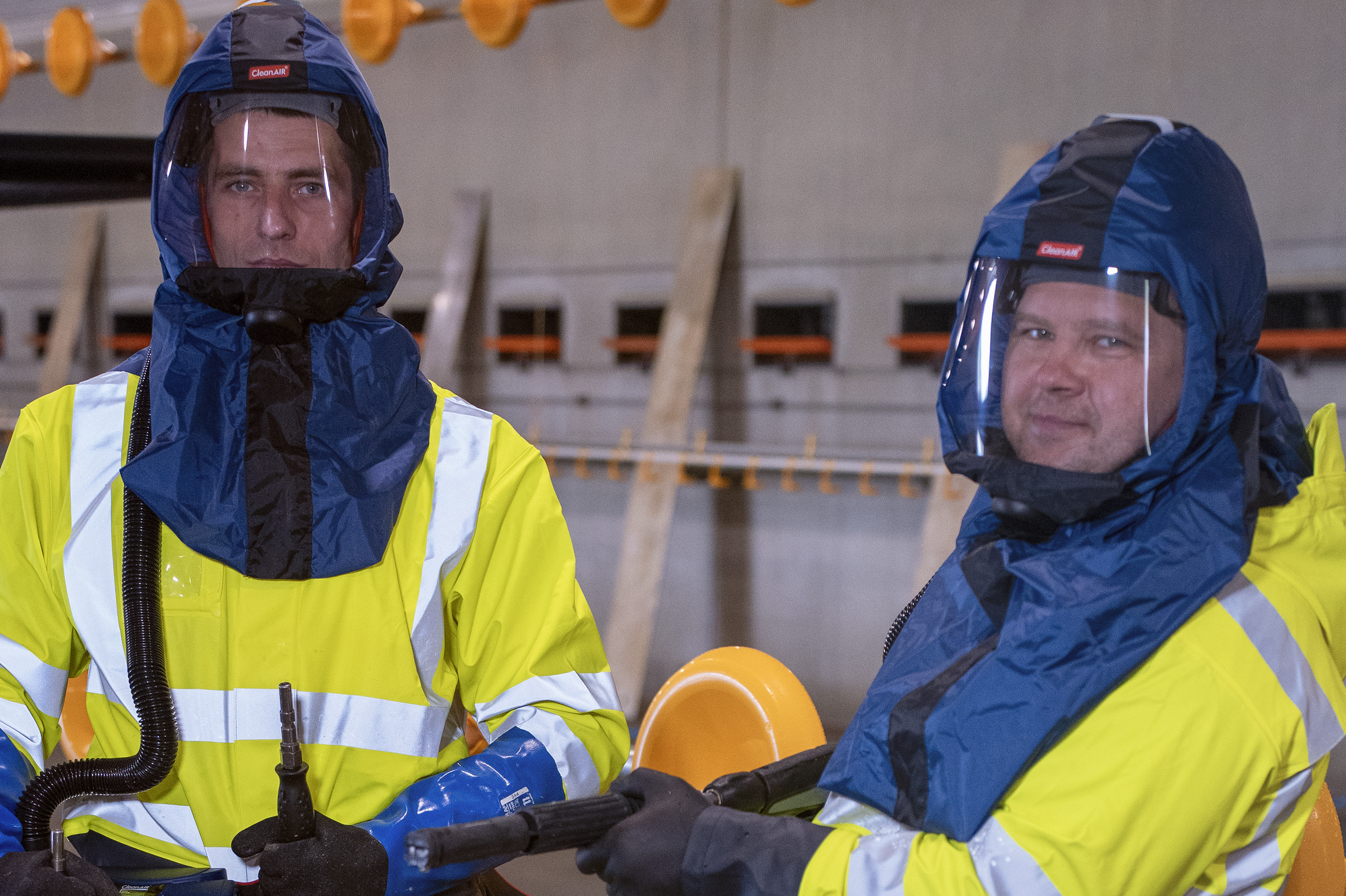 To personer iført høye synlige våtdrakter og visir står i en indendørs industriatmosfære.