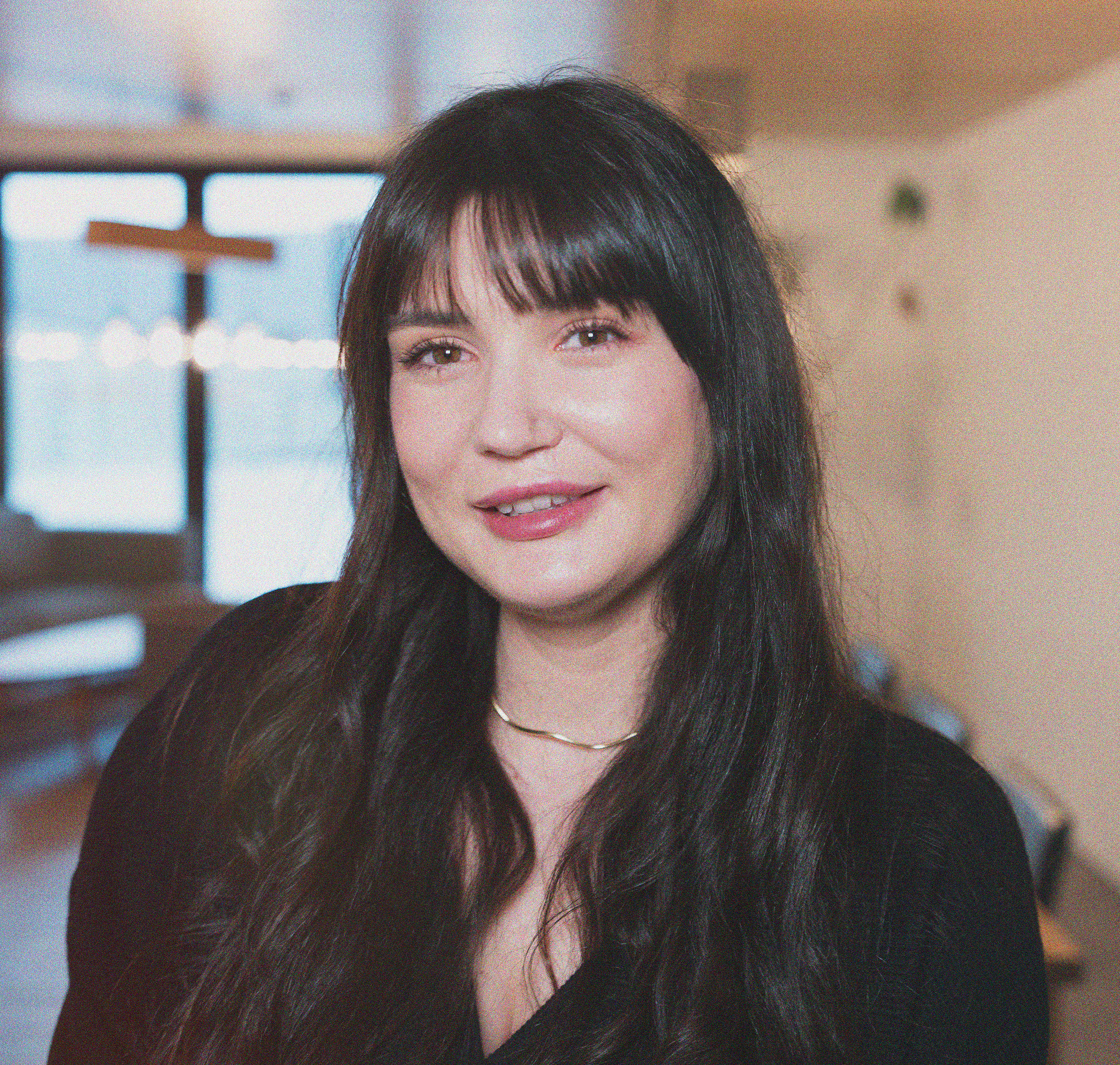 A woman with long, wavy hair and bangs smiling at the camera, wearing a dark blazer over a striped shirt, against a plain background.